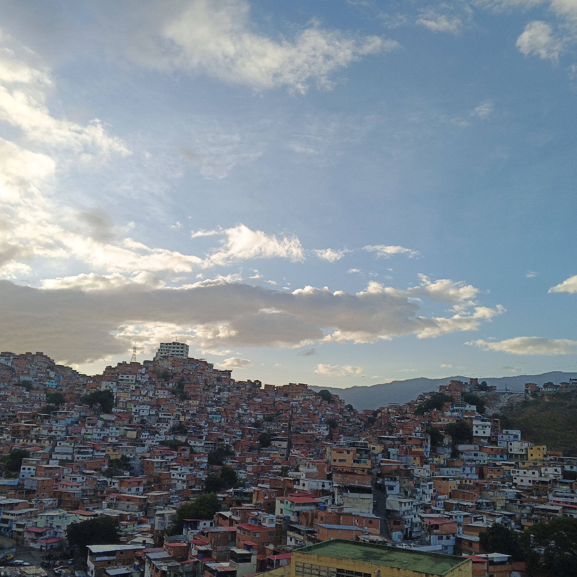 An expansive, high-angle view captures a densely populated urban hillside, characteristic of a favela or barrio in Caracas, Venezuela, under a partly cloudy sky. The scene is dominated by countless small, multi-story buildings sprawling across undulating terrain, with distant mountains forming the horizon.
The architecture is informal, with houses tightly packed together, displaying a mix of exposed red brick, concrete, and facades painted in various colors such as white, light blue, green, and orange. Many roofs appear to be flat or slightly sloped, often made of corrugated metal or concrete. On a prominent peak within the settlement, a taller, white, multi-story building stands out, accompanied by a communication tower. Green foliage from scattered trees is visible between the structures, providing small breaks in the dense construction. A few indistinct vehicles can be seen in the lower left portion.
The sky occupies the upper half of the image, showcasing a bright blue expanse dotted with white and grey cumulus clouds. The lighting suggests late afternoon or early evening, with warm sunlight illuminating the clouds from the side and casting a soft glow over parts of the landscape. The weather is partly cloudy. No specific activity or event is taking place, depicting a static landscape view. No people or discernible text are visible in the image.
