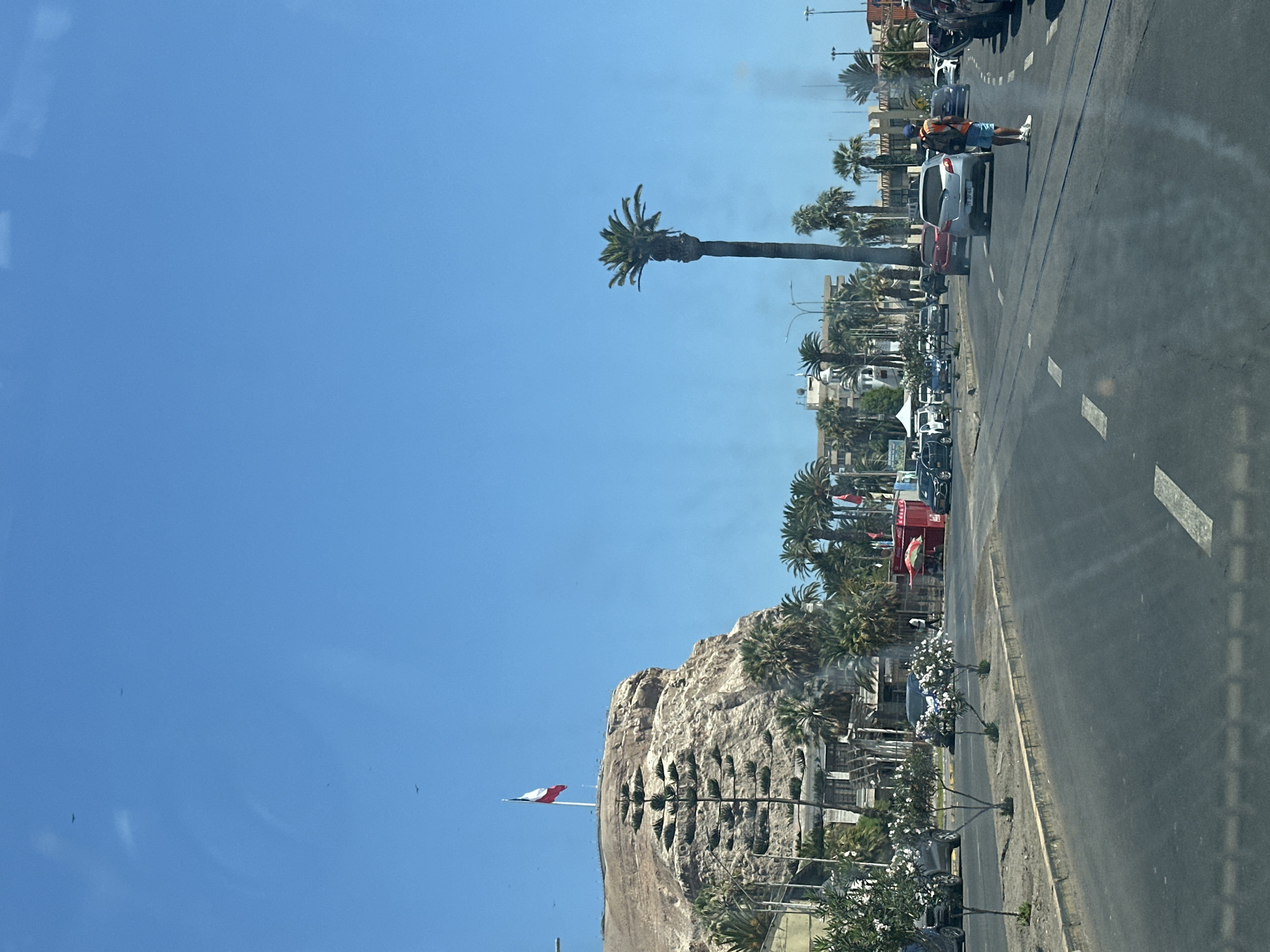 View from a vehicle traveling on a road in Arica, Chile. The image captures a cityscape with palm trees lining the street. A distinctive geological feature, likely El Morro de Arica, is visible, adorned with a large Chilean flag. Traffic is present on the road with pedestrians. The sky is clear and blue.