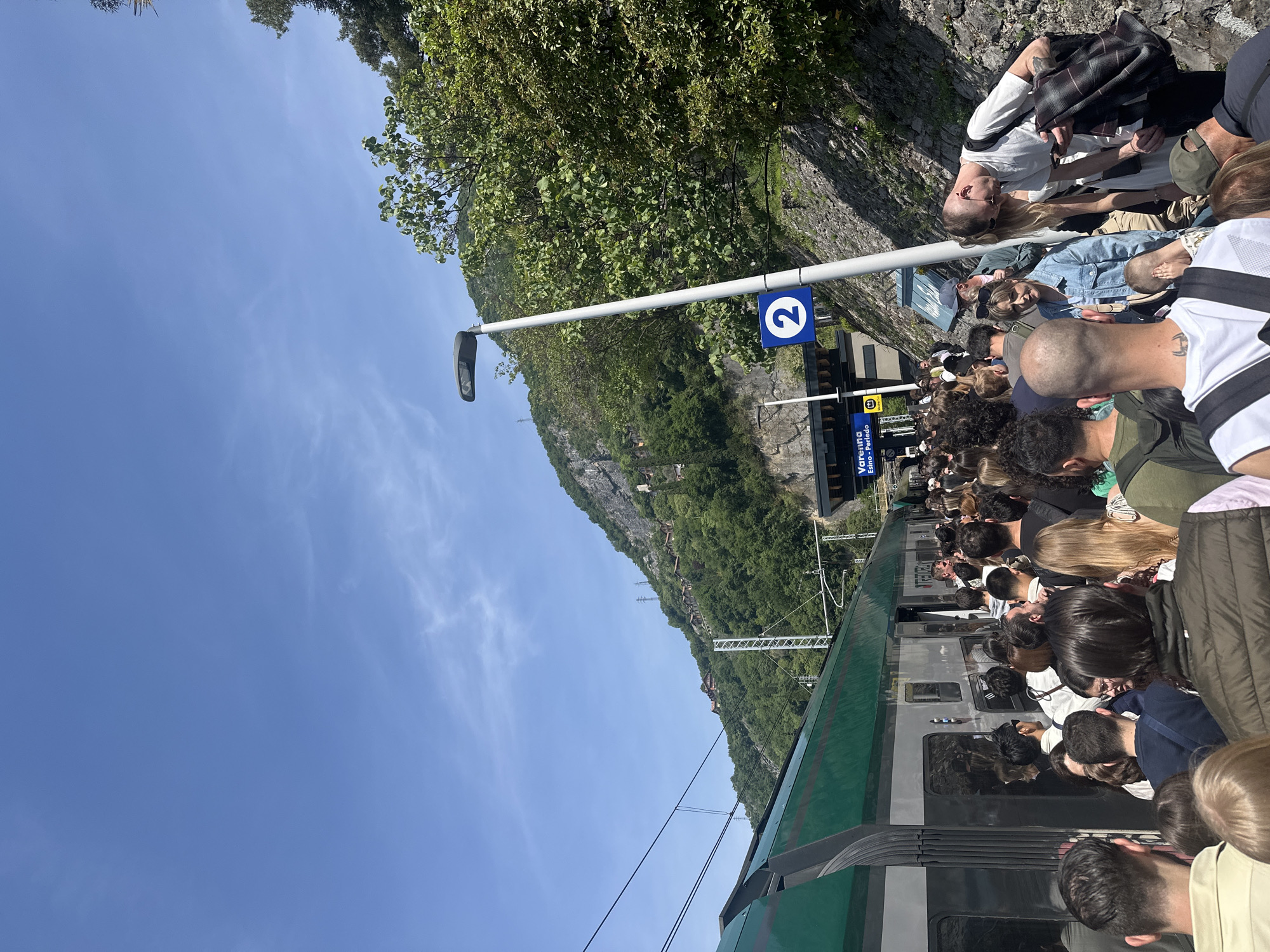 The image depicts a crowded train platform at the Varenna-Esino-Perledo train station in Gittana, Italy. Passengers are densely packed near the train doors, presumably boarding or disembarking. A green and silver train is visible alongside the platform. A sign indicates the platform number '2'. Beyond the platform, a steep hillside with vegetation and some visible structures rises into the background. The sky is clear and blue. A light pole stands near the platform edge, and a stone wall runs along the upper part of the scene. Several people are seen standing or sitting near the wall.