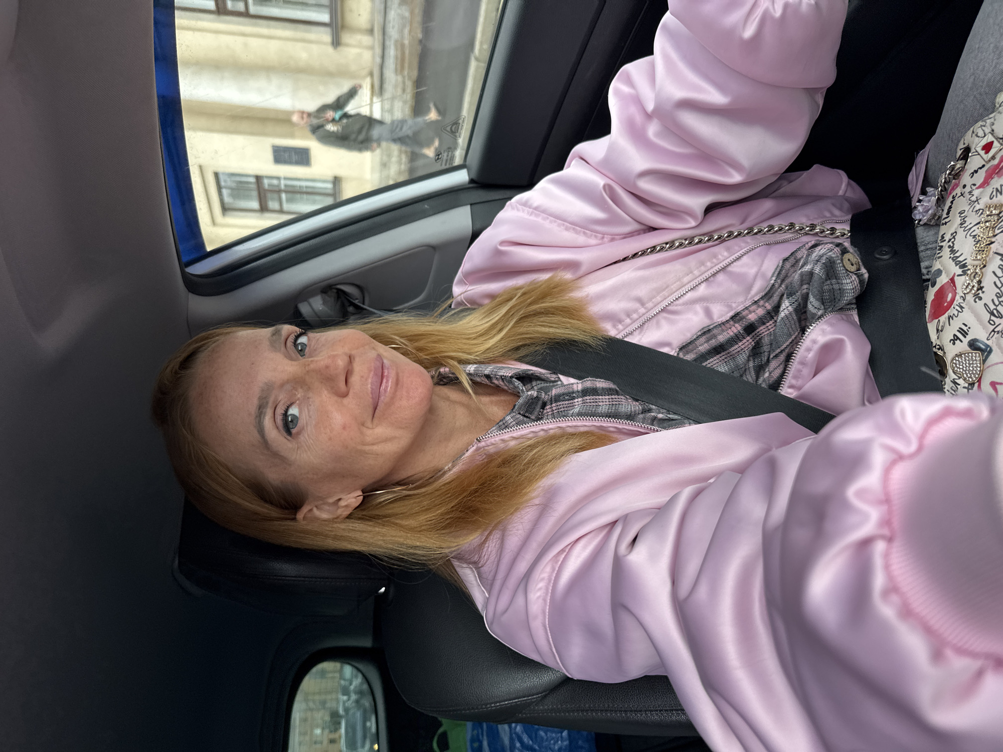 A blonde woman in her late 40s or early 50s is seated in the driver's seat of a car, looking towards the camera. She has fair skin, freckles, and light blue eyes. She is wearing a pink satin bomber jacket over a plaid shirt and a light-colored purse with a chain strap rests on her lap. The car's interior is visible, including a black leather seat and a seatbelt. Through the car window, a blurred background shows buildings and a pedestrian walking with a cane on a street. This scene is likely in Saint Petersburg, Russia, given the architectural style visible through the window and the overcast weather conditions often associated with the city. The woman appears to be holding a phone, suggesting she might be taking a selfie or recording a video. The overall impression is a candid shot taken during a car ride, possibly in a city environment.
