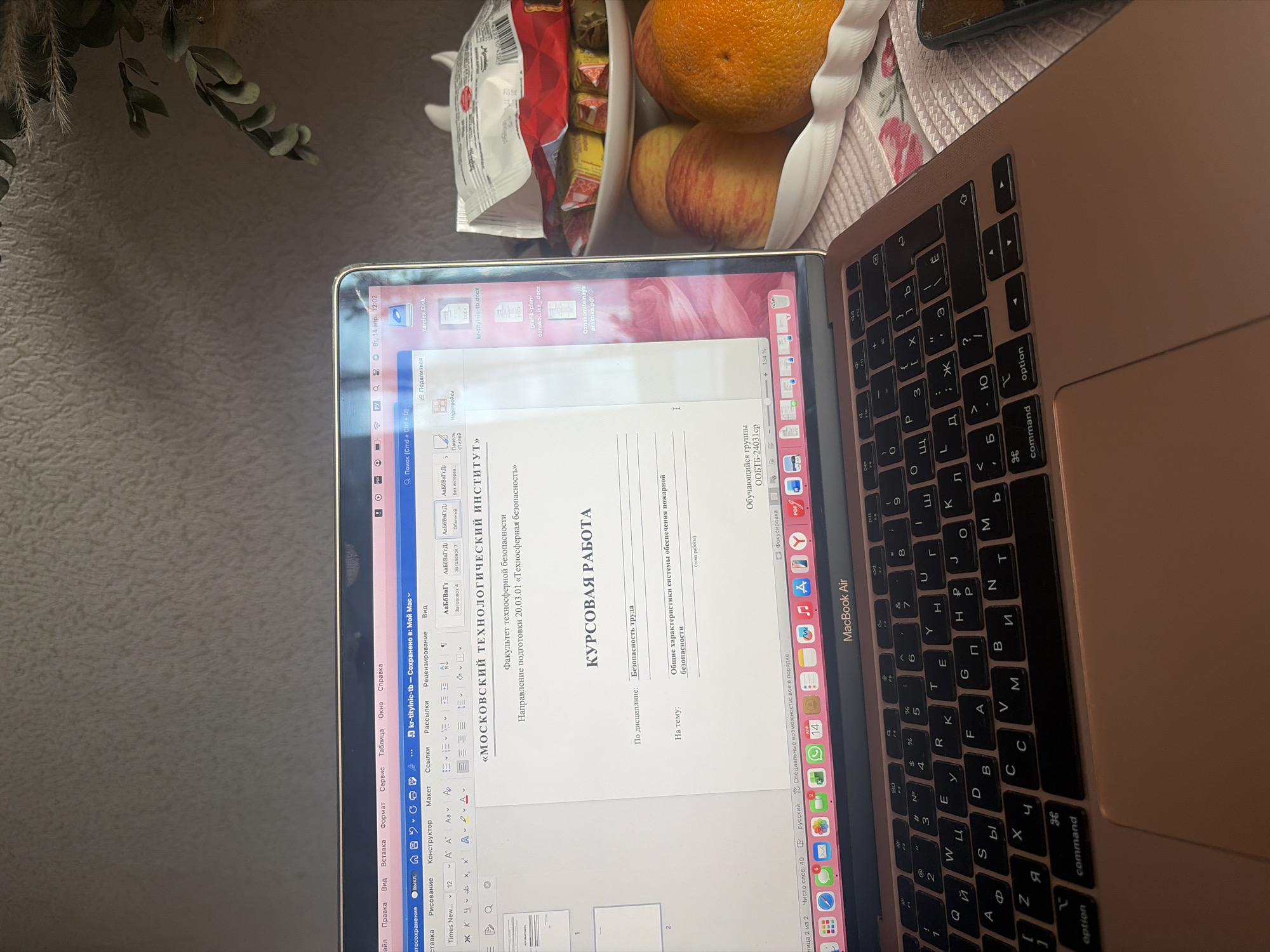 The image shows a MacBook Air open on a table. The screen displays a Word document in Russian, titled "COURSEWORK" with the header "MOSCOW TECHNOLOGICAL INSTITUTE" visible. The laptop keyboard has Russian characters. To the side of the laptop, on the table, there is a white bowl containing fruit (an orange and apples) and a package of sweets. Part of a mobile phone is visible in the upper right corner. The background is blurred, showing plant stems and leaves.