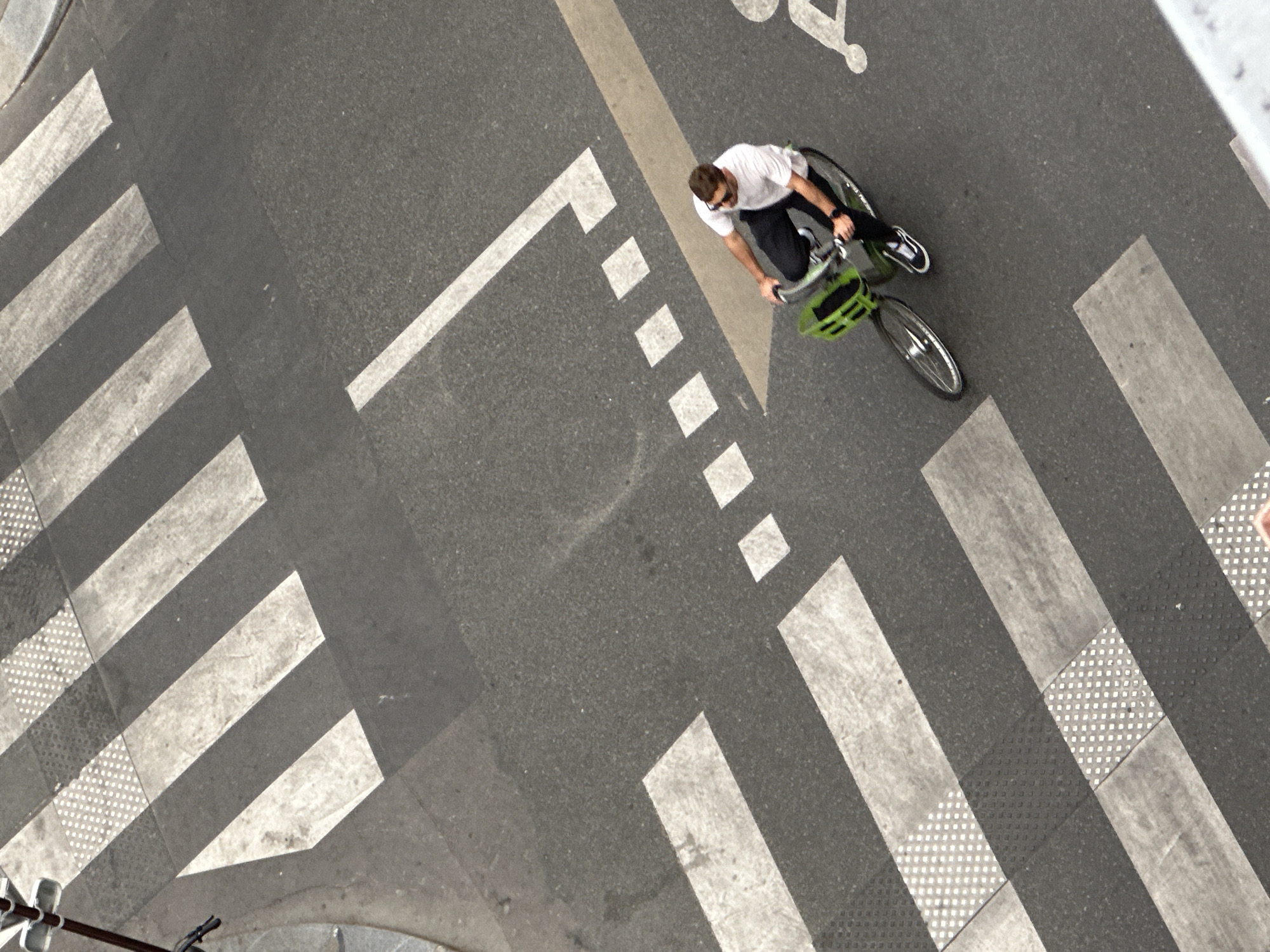 An overhead, high-angle shot captures a single cyclist navigating an urban street. The scene is dominated by dark gray asphalt marked with various white road markings, suggesting an intersection or a dedicated crossing area for pedestrians and bicycles.
The main subject is a person on a bicycle, viewed from directly above. The cyclist wears a white short-sleeved t-shirt, dark trousers, and sunglasses. Their legs are actively engaged in pedaling, with one foot visible in a white and black sneaker. The bicycle itself is dark-colored and features a distinctive bright green basket mounted on the front handlebars.
The road surface displays an intricate pattern of white markings. On the left side of the frame, a prominent pedestrian crosswalk consists of multiple thick, parallel white stripes. Some sections of these stripes, particularly towards the bottom left and top right, include a textured, dotted pattern. Extending from the bottom-left towards the center, there is a bold, solid white L-shaped marking, followed by a series of evenly spaced white square dashes. To the right of the cyclist, a distinct solid white line runs diagonally across the asphalt, likely delineating a specific lane or boundary. Further in the upper right quadrant, a stylized white bicycle symbol is painted on the road, clearly indicating a bicycle lane or path, consistent with an urban European setting like Paris.
The lighting suggests bright daytime conditions, with clear visibility and no harsh shadows, implying either direct sunlight or a brightly overcast sky. The image provides a clean, clear view of the cyclist and the surrounding street infrastructure.