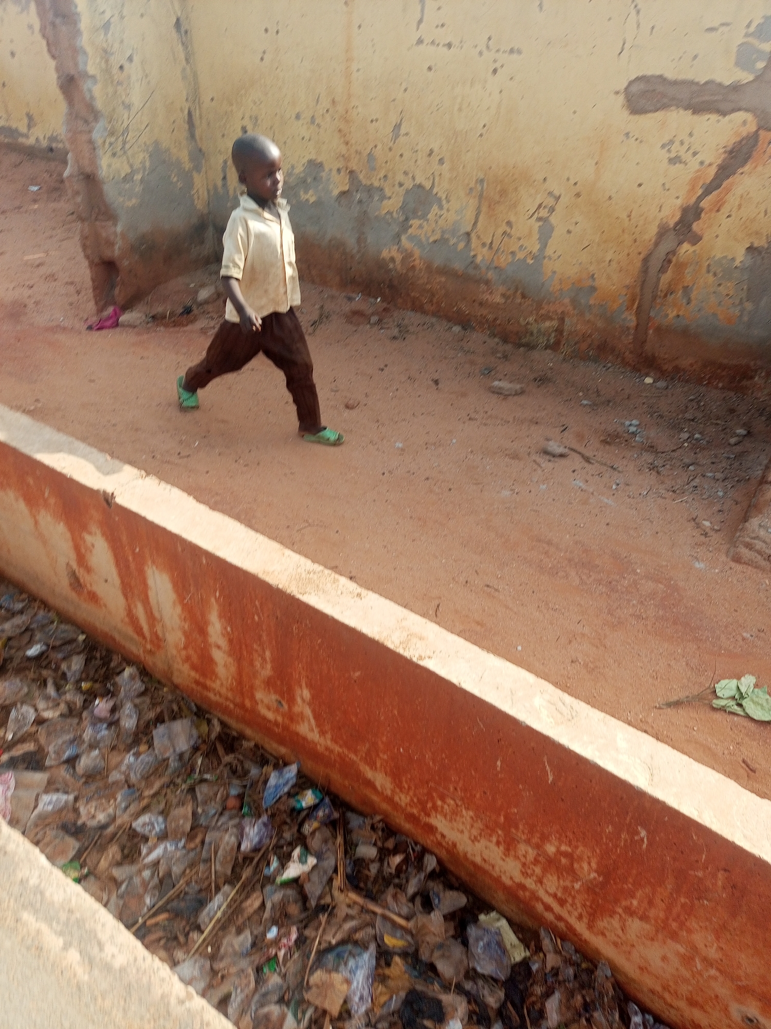 In a daytime shot taken in Gombe, Nigeria, a young child is walking on a dirt road adjacent to a concrete drainage ditch filled with litter. The child is wearing a light-colored shirt, brown pants, and green sandals. The background features a weathered wall with peeling paint. The location appears to be a poorer, less developed area of the city.
