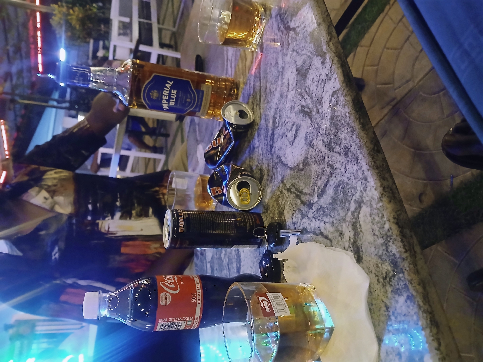 The image shows a bar or restaurant table in Jos, Nigeria, littered with alcoholic beverages and associated items. There is a partially full bottle of Imperial Blue whiskey, two empty and crushed cans of Red Bull, a bottle of Coca-Cola, two glasses containing amber-colored liquid, likely alcoholic drinks. A set of car keys is placed amongst the clutter on a white napkin or tissue. A person is partially visible on the left side of the image, wearing a patterned shirt. The table appears to be made of marble or a similar stone material. In the background are out-of-focus lights and what appear to be chairs, suggesting an outdoor or semi-outdoor setting.