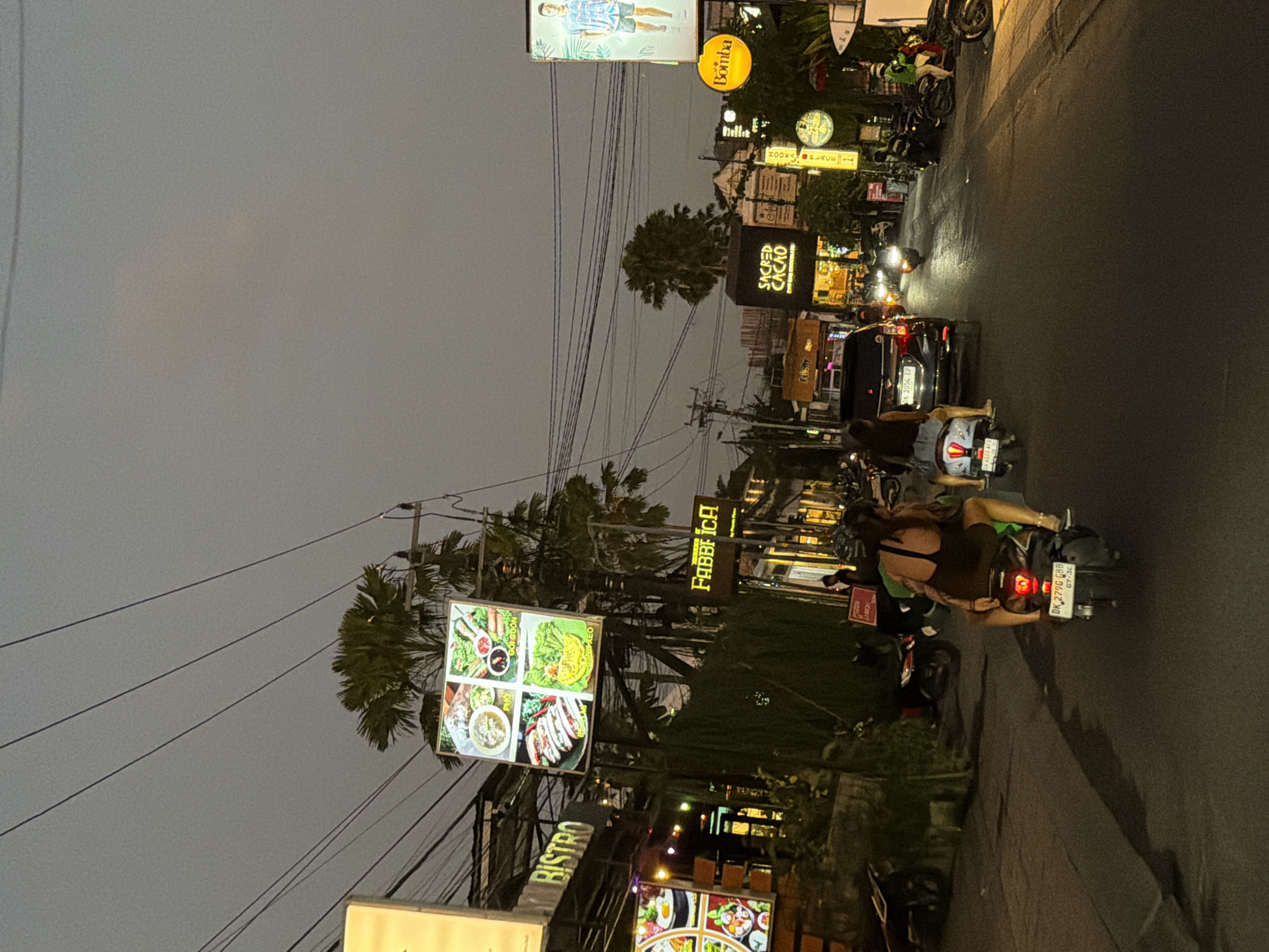 This image captures an evening street scene in North Kuta, Bali, Indonesia, characterized by active traffic and illuminated commercial establishments.
In the immediate foreground, the back of a person riding a black Honda scooter is visible, with the license plate "DK 2796 GBB" partially legible. This individual wears a dark top and shorts. Slightly ahead and to the left, another person on a lighter-colored scooter, bearing the license plate "DK 6573 AFG," is also facing away from the camera. A black car, identified by its license plate "DK 1054 EP," is situated between these two scooters.
The asphalt street extends into the midground, where additional motorcycles and at least one car are seen, their headlights and taillights glowing. On the left side of the street, multiple businesses are visible. An illuminated sign for "Bistro" is prominent, accompanied by a large, bright sign displaying a four-quadrant food menu featuring dishes like "Pho," "Goi Cuon," "Cha Gio," and "Banh Xeo." Further down, another illuminated sign reads "FABBRI CA." Tropical trees and dense green foliage line the street, partially obscuring some building facades.
On the right side of the street, an illuminated sign for "SACRED CACAO" is clearly visible, with its building appearing dark. Further along, a circular yellow sign for "Bomba" and a vertical sign for "HOORA PLACE" contribute to the vibrant street lighting. A tall billboard on the far right displays a male model in a casual shirt and shorts.
The sky above is dark grey and overcast, indicating dusk or early night. Numerous dark power lines crisscross the upper portion of the frame, supported by utility poles visible on both sides of the street. The overall environment depicts a bustling urban street with diverse commercial activity under artificial lighting.