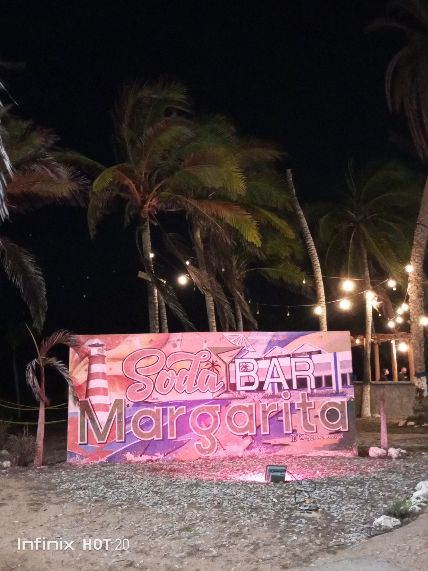 This nighttime photograph, taken in Porlamar, Venezuela, showcases a vibrant outdoor scene dominated by a large, brightly lit sign. The sign, centrally positioned, prominently displays the text "Soda BAR Margarita" in colorful, stylized fonts of pink, orange, and purple. It features a decorative graphic with a lighthouse on the left and what appears to be a coastal town silhouette on the right. A ground-level spotlight illuminates the sign with a distinct pinkish-purple glow, casting light onto the unpaved ground covered with gravel and small rocks in the foreground.
Numerous tall palm trees frame the scene, their silhouettes visible against the dark night sky. Several strands of warm, glowing string lights are strung between the palm trunks, providing ambient illumination and creating an inviting atmosphere. To the far right, a rustic wooden structure, possibly part of a bar or seating area, can be seen, with two vague figures of people discernible within it, adding to the sense of an active social space.
The image conveys a relaxed, tropical evening ambiance, typical of a bar or restaurant setting. The time of day is clearly night, as indicated by the dark sky and the reliance on artificial lighting. A "Infinix HOT 20" watermark is visible in the bottom left corner, identifying the camera. Below the "Margarita" text on the sign, a smaller social media handle, likely "@SODABARMGTA," is also visible.