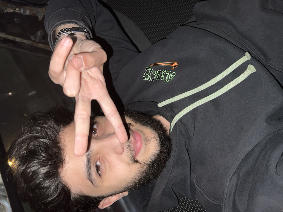 A young man with dark curly hair and a beard is posing for the camera. He's wearing a black hoodie with a Nike logo in green and orange. He is flashing the peace sign. He has a silver bracelet on his wrist. The background appears to be the interior of a car.