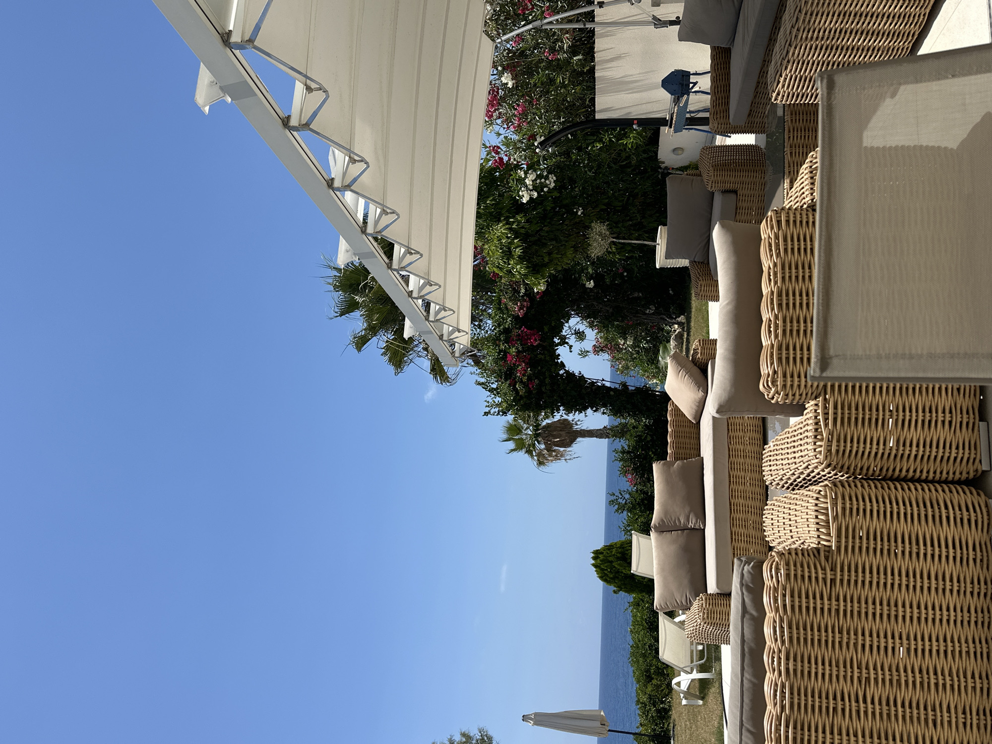 This is a wide outdoor shot taken from a ground-level perspective, showcasing a patio area with rattan furniture and a view of the sea. In the foreground, a collection of plush, tan-cushioned rattan sofas and armchairs are arranged, some with woven detailing. Behind the furniture, a lush green lawn leads to a white retractable awning supported by a metal frame. Beyond the lawn, the deep blue Mediterranean Sea is visible under a clear, bright blue sky. A few palm trees dot the landscape, and a white patio umbrella stands closed on the left. A stone archway covered in vibrant pink and white flowers frames the view of the sea and foliage. The scene appears to be a relaxation or dining area at a resort or private residence in the Kissonerga Community, Cyprus, given the typical climate and vegetation suggested by the image. There are no people or animals visible in the frame.