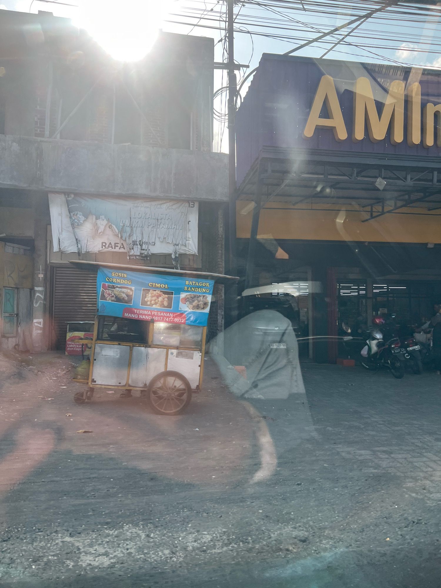 The image shows a street scene in Payaman, Indonesia. In the foreground, a food cart is visible, selling "Sosis Corndog," "Cimol," and "Batagor Bandung." The cart has a blue banner with images of the food items and text that reads "TERIMA PESANAN MANG MANG 0817 7417-0032." Behind the food cart, there is a building with a sign that partially reads "AM". Further back, several motorcycles are parked in front of a building with a yellow awning. The sun is shining brightly, creating lens flare and reflections in the image.