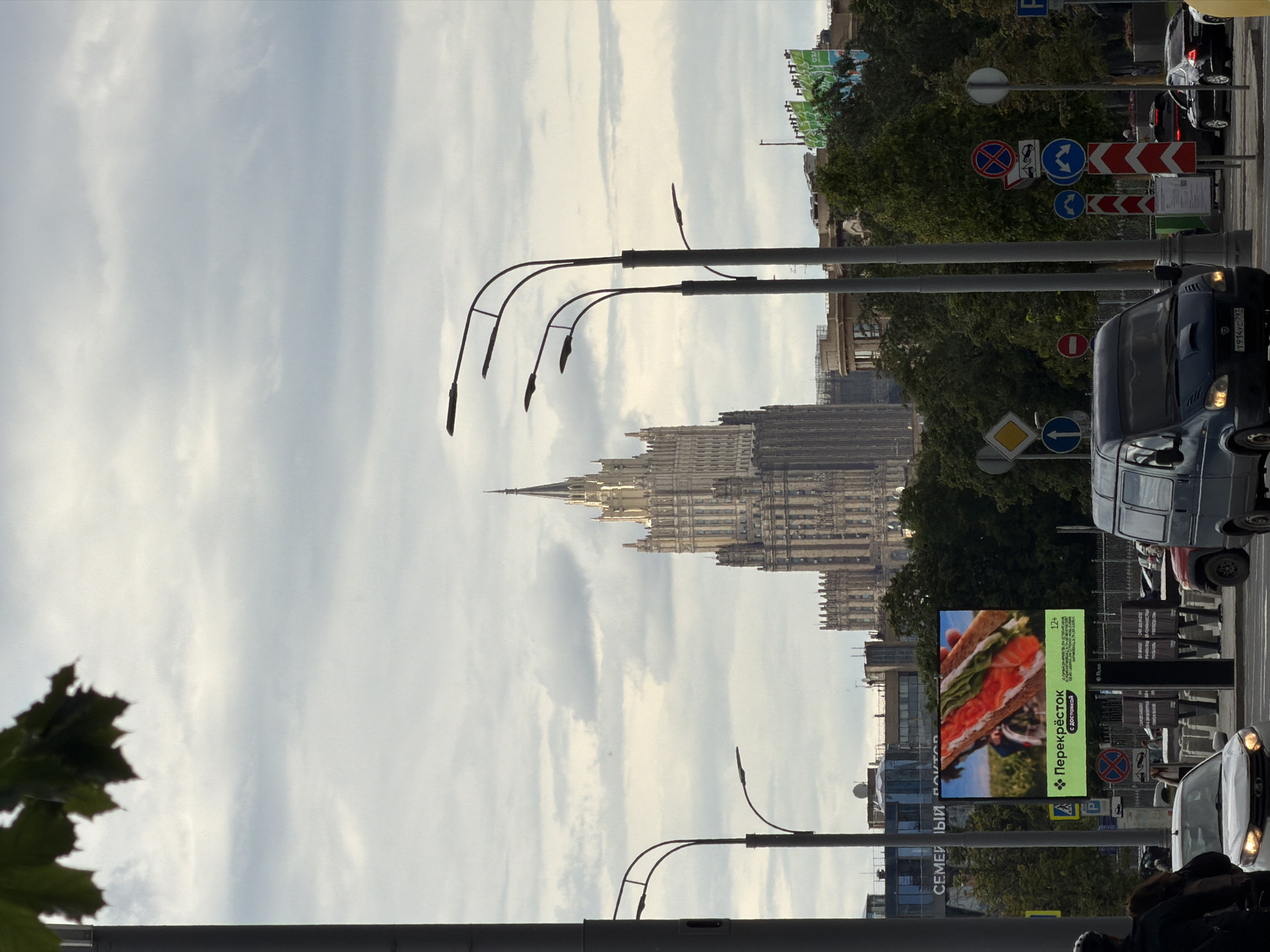 The image depicts a cityscape in Moscow, Russia. The Ministry of Foreign Affairs building, one of the Seven Sisters skyscrapers, is visible in the background. Streetlights line the road, and several vehicles, including a dark van with license plate Y914XC797, are seen in motion. Traffic and regulatory signs are posted along the street. A large digital billboard displays an advertisement for a food product with the word "Перекрёсток" and other text in Cyrillic. The sky is overcast. The scene suggests a busy urban street with a mixture of historical architecture and contemporary commercial elements.