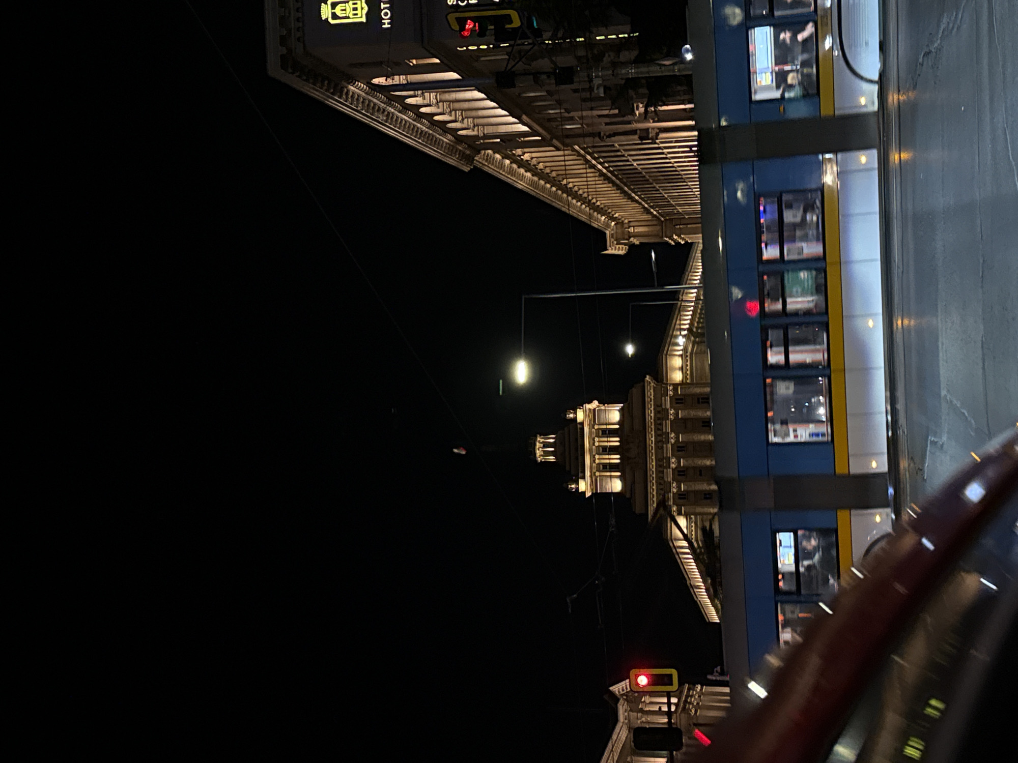 The image captures a night scene in Sofia, Bulgaria. A tram with blue and white livery is passing by, illuminated by the streetlights. The architectural details of a building, possibly a hotel, are visible in the background. A red traffic light is active. The perspective suggests the photo was taken from a vehicle. The sky is dark, emphasizing the city's artificial lighting.