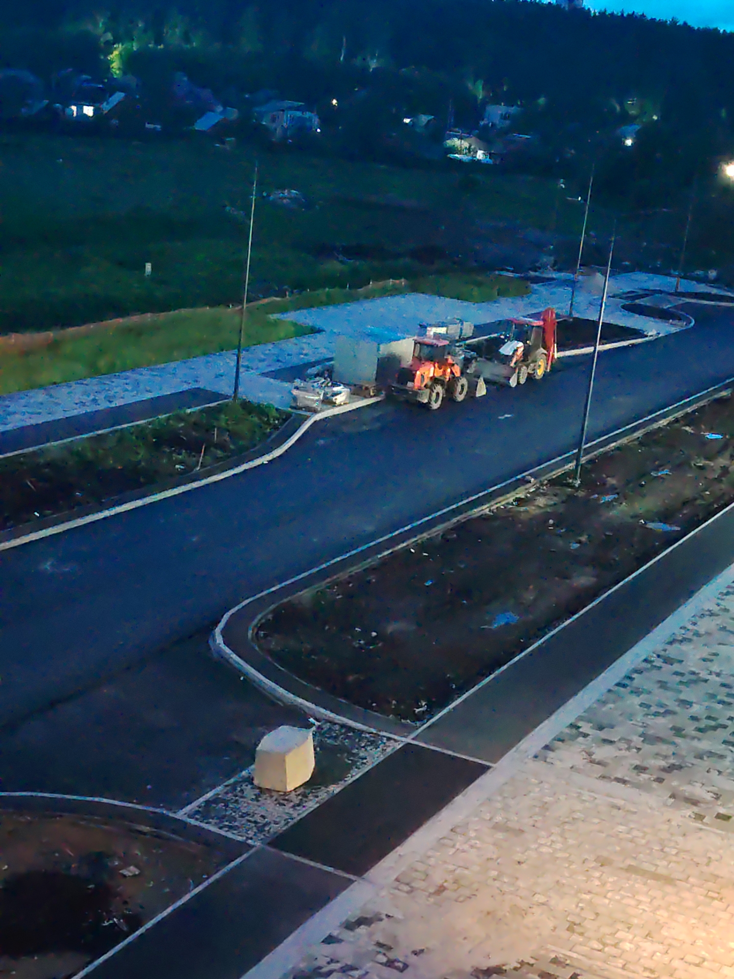 The image shows a construction site at dusk in Yekaterinburg, Russia. Two construction vehicles, an orange tractor and a red backhoe, are parked on a paved road. In the background, several houses are visible on a hillside, with trees and a dark sky above. The foreground features a paved walkway and a light-colored, cubic object. The scene suggests ongoing development and infrastructure work.