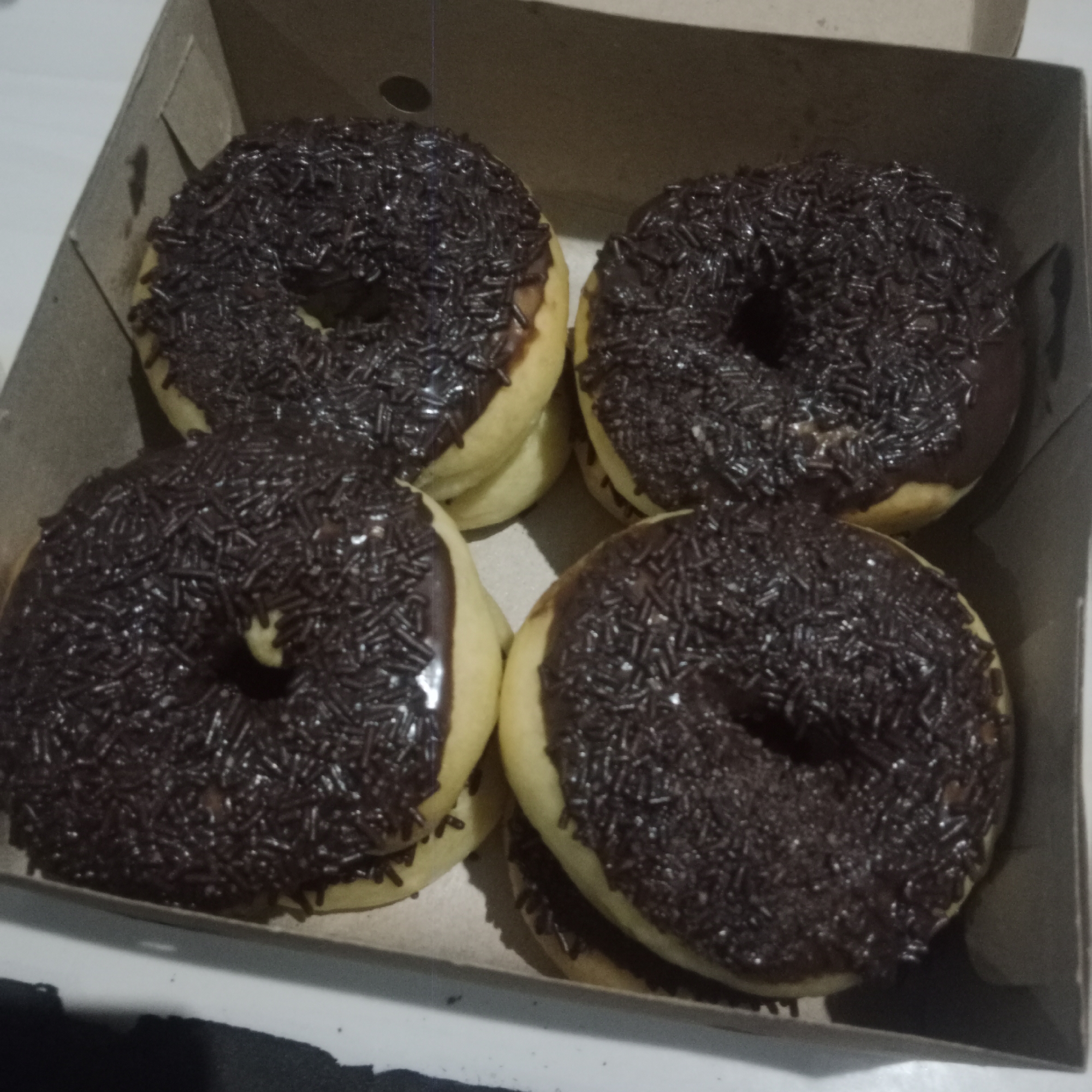 Four chocolate-frosted donuts with chocolate sprinkles are arranged in a brown cardboard box. The donuts are circular with a hole in the center. They are positioned in a 2x2 grid within the box. The box appears to be made of corrugated cardboard and has slightly open flaps. The background suggests an indoor setting, possibly a table or countertop, with a white surface visible. There are no people present in the image. The scene depicts a simple food item presentation. The location is Tegowanu Kulon, Indonesia.