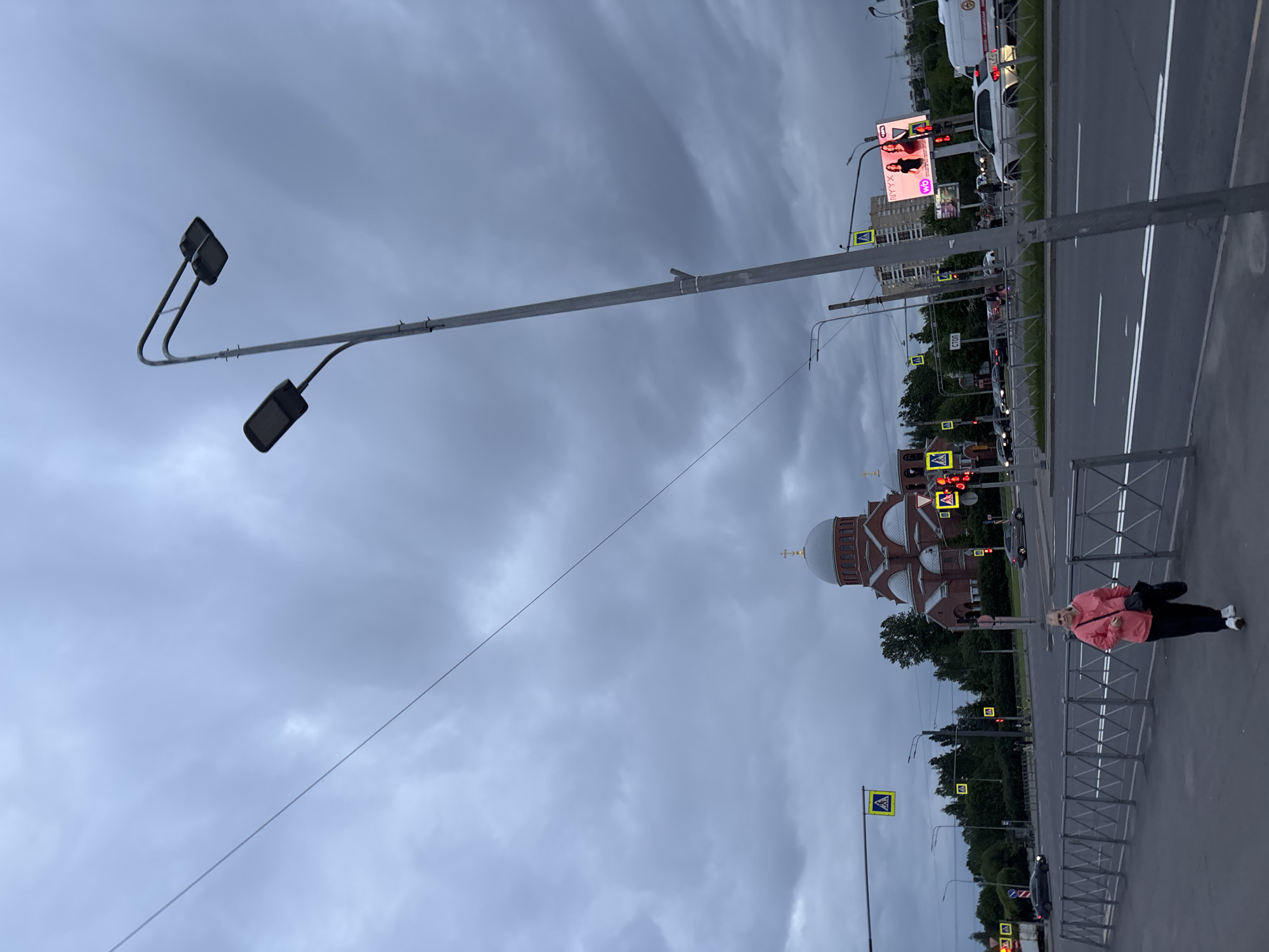 The image depicts a street scene in Unknown. A church with a white dome and multiple crosses is visible in the background. A woman in a pink jacket and black pants is walking on a sidewalk next to a metal railing. A street runs parallel to the sidewalk, with cars and traffic lights. Streetlights are positioned along the sidewalk. The sky is overcast.