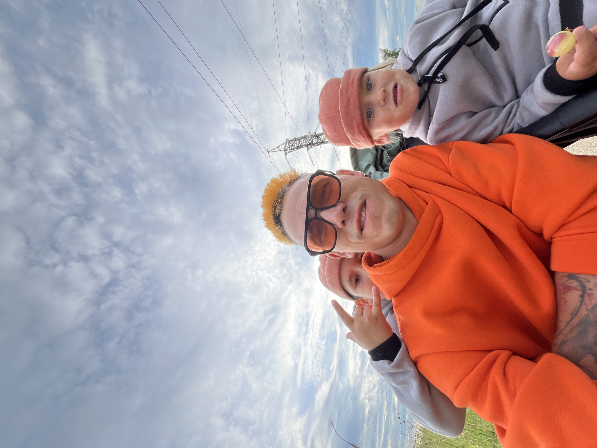 In this selfie-style image, three individuals are positioned against a cloudy sky with visible power lines and a distant power tower, suggesting an outdoor, possibly industrial or rural setting. The person in the foreground, wearing an orange hoodie and sunglasses, has brightly dyed orange hair and is smiling at the camera. To their left and slightly behind, another person, a child wearing a peach-colored beanie, looks directly at the camera with a surprised expression. A third individual, mostly obscured, is wearing a grey hoodie and is seen holding a small, colorful object. The lighting indicates it's daytime, with overcast conditions. The overall mood appears lighthearted and casual, as suggested by the smiling faces and the peace sign gesture made by the child. The image is taken in Chaikovsky, Russia. There is no visible text in the image.