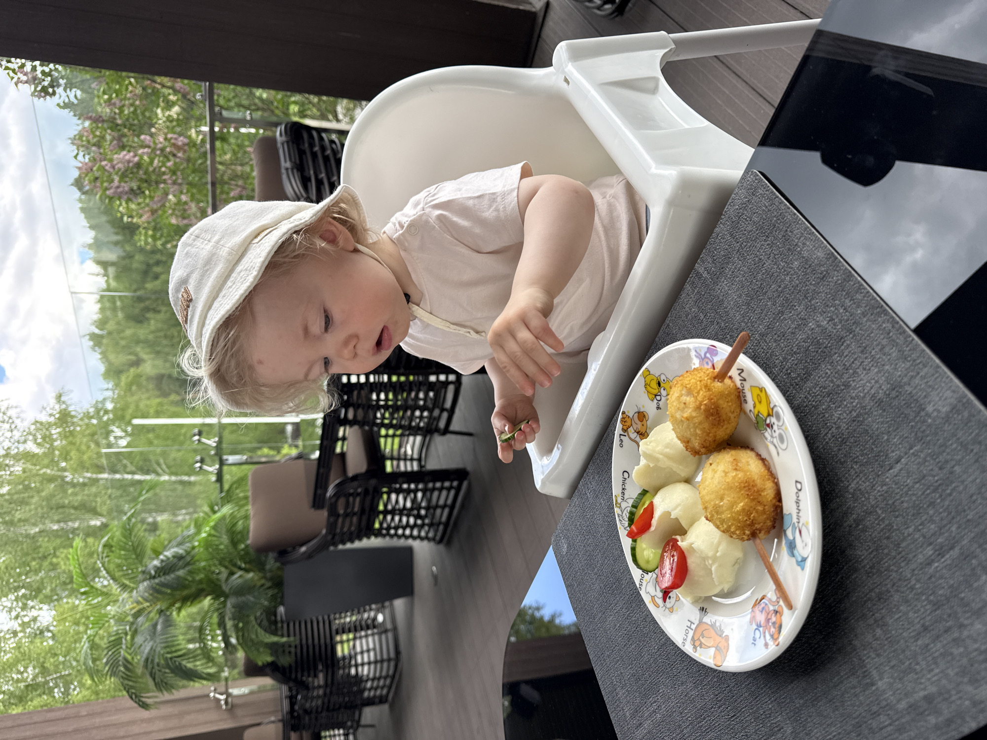 The image shows a young child in a white high chair on an outdoor terrace or balcony during daytime. The child, with light hair and wearing a light t-shirt and a cream bucket hat, is looking down at a plate of food placed on a dark grey placemat. In their left hand, the child holds a small piece of green food, likely cucumber.
The white plate features colorful cartoon character illustrations and names around its rim, including "Leo," "Mouse," "Chicken," "Dolphin," "Cat," "Horse," and "Doraemon." The meal consists of two golden-brown, breaded spherical items on wooden skewers, served alongside scoops of mashed potatoes and a small salad of sliced cucumber and cherry tomatoes. Adjacent to the child's eating area is a dark, reflective table surface.
The background reveals a vibrant outdoor setting with green trees and a cloudy sky visible through a glass railing. Dark wicker patio furniture is also discernible. The overall scene suggests a calm outdoor meal.