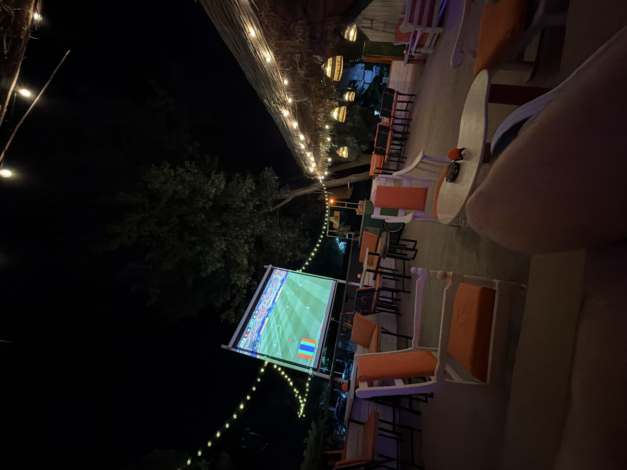 The image shows an outdoor seating area at night, likely a cafe or bar, with a large projector screen displaying a soccer match. The area is decorated with string lights and thatched roofing. There are several tables and chairs, most of which are orange and white. The ground is paved. In the foreground, a person's leg is visible, suggesting the photo was taken from a seated position. The scene is set in Tsarevo, Bulgaria, as indicated by the context.