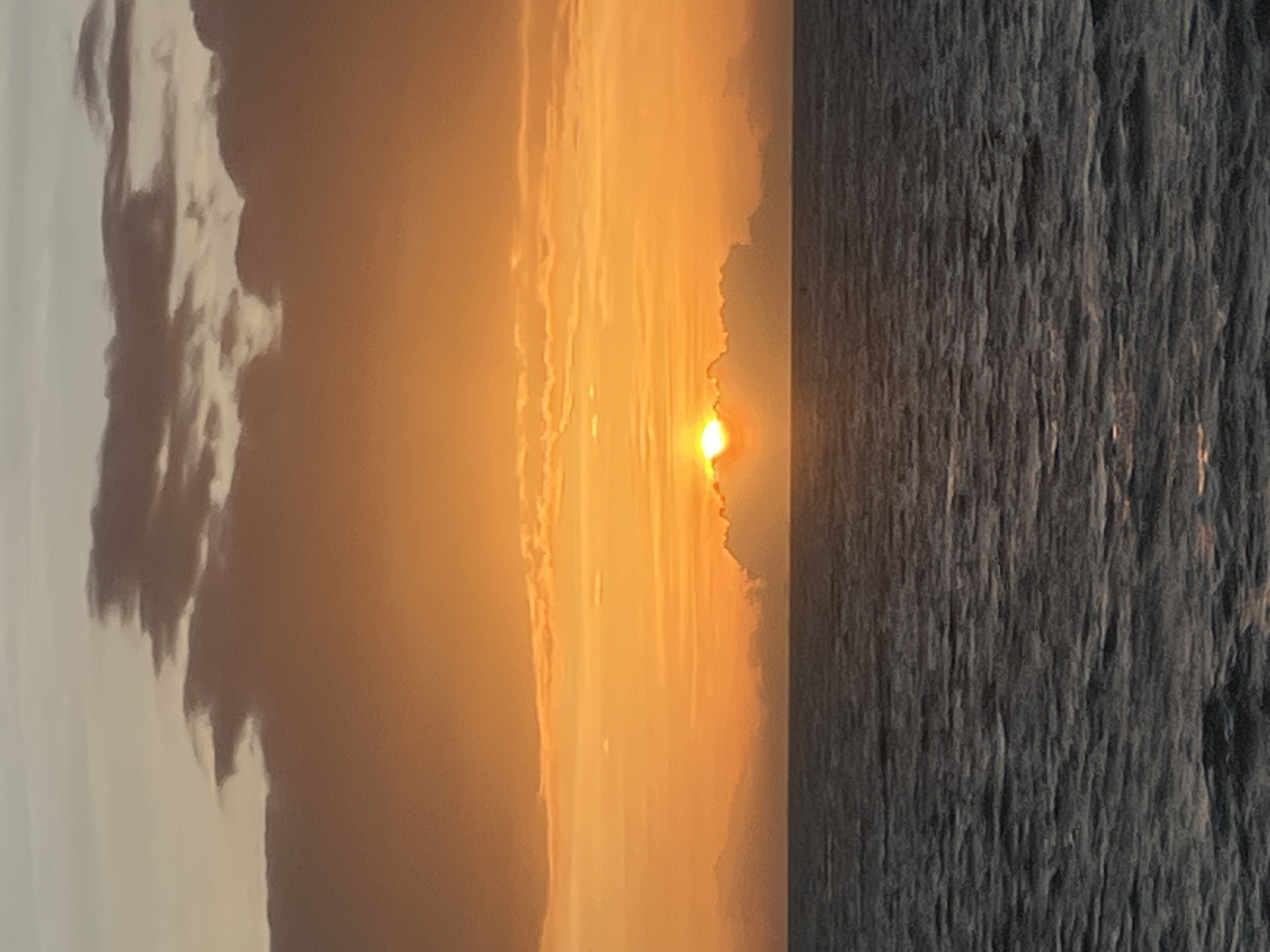 This is a photograph of a sunset over the ocean, likely taken from a beach or boat. The sky is a vibrant gradient of orange and yellow, with clouds scattered across it. The sun is a bright orb peeking through the clouds near the horizon. The ocean is calm and dark, with gentle waves reflecting the colors of the sky. There are no people or man-made structures visible in the image, suggesting a natural and serene setting. Given the location of Ko Pha-ngan District, Thailand, this scene is likely depicting a beautiful sunset over the Gulf of Thailand.