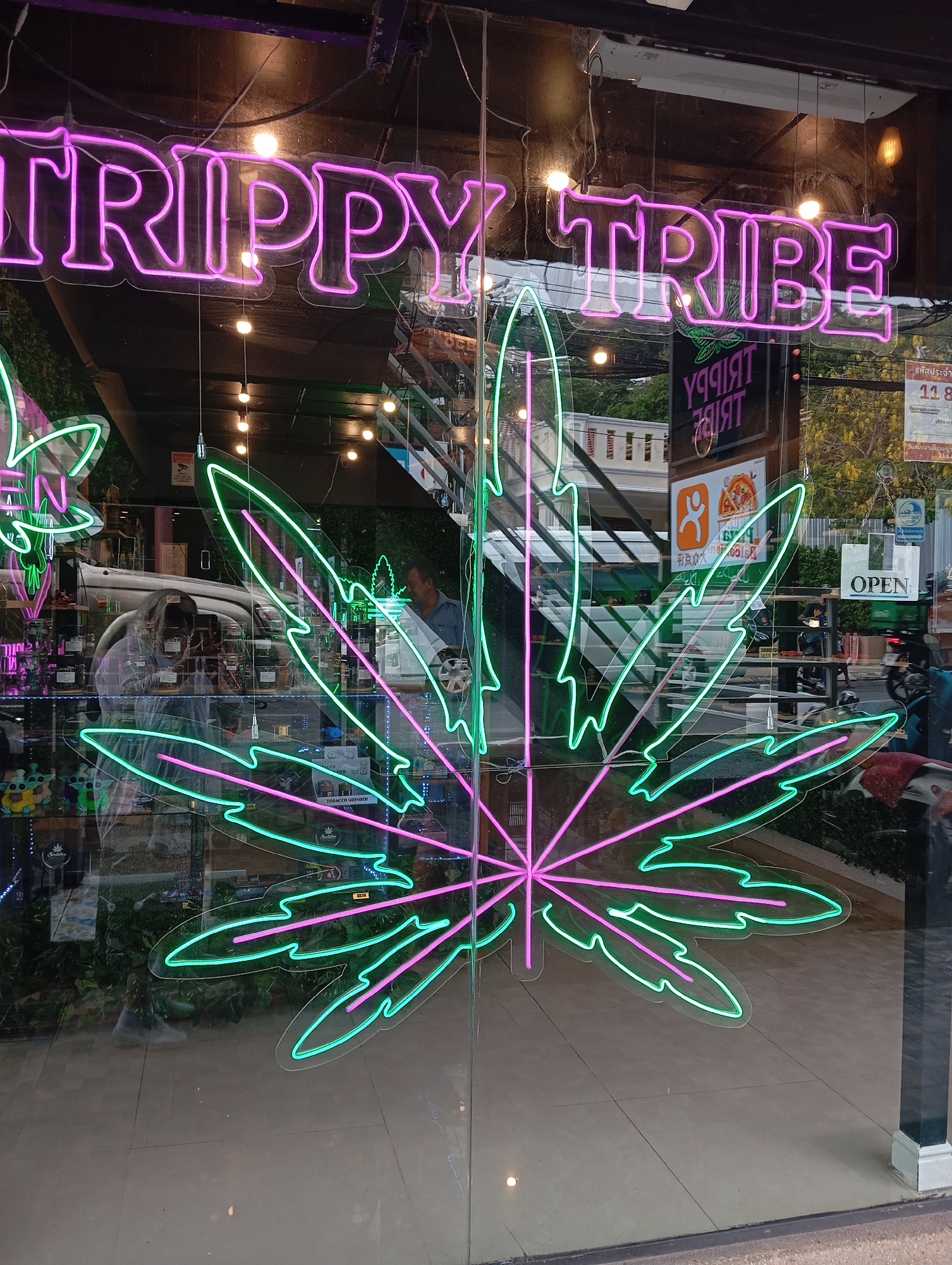 The image is a close-up shot of a storefront in Karon, Thailand, featuring vibrant neon signs reflected on the glass. A large, stylized neon cannabis leaf, rendered in shades of green and pink, dominates the center of the frame. Above it, a neon sign in hot pink reads "TRIPPY TRIBE". Reflected in the glass are elements of the street outside, including buildings, trees, a motorcycle, and a person. Inside the store, shelves stocked with products are visible, along with a security camera. Another neon sign inside the store appears to be an inverted version of "TRIPPY TRIBE". A sign on the right reads "OPEN". The floor inside the store is tiled.