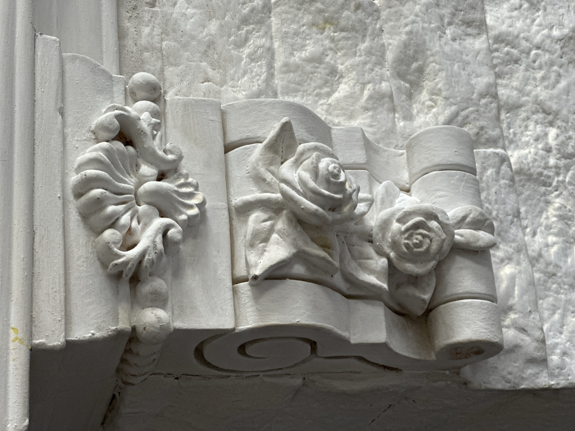 A white, ornate architectural element is attached to a white brick wall. The element is a corbel featuring carved floral motifs, specifically roses and acanthus leaves. The corbel is composed of plaster or a similar material and is painted white. The brick wall behind it is textured and appears to be painted or whitewashed white as well. The lighting suggests the image was taken indoors, possibly in a historical building or a room with decorative architectural features. There are no people or animals present in the image.