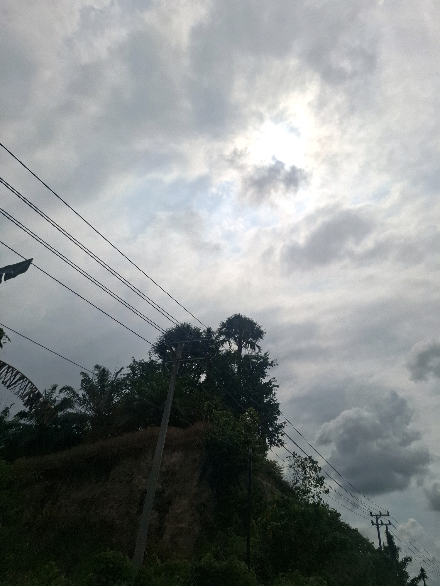 The image depicts an overcast sky with the sun peeking through dense clouds, creating a hazy luminescence. In the foreground, power lines stretch diagonally across the frame from the upper left to the lower right. Below the power lines, a steep, eroded hillside is covered in dense green vegetation, including trees with palm-like fronds. A single utility pole stands on the hillside, with wires attached. The overall impression is of a natural landscape in Seuneubok, Indonesia, with man-made infrastructure present. The scene appears to be captured during daylight, possibly in the late afternoon or early morning due to the diffused sunlight. There are no people or animals visible in the image.