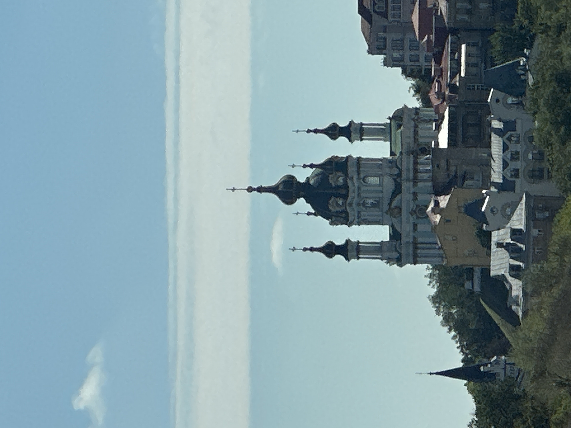 This is a photograph of St. Andrew's Church in Kyiv, Ukraine. The church is a Baroque-style architectural monument located on the steep Andriyivskyy Descent. It is depicted from a distance, with the church's iconic blue domes and white facade dominating the central part of the image. The sky above is a clear blue with some faint clouds. In the foreground, there are rooftops of other buildings and lush green trees, suggesting a hilly urban landscape. The overall impression is of a picturesque and serene view of a historic landmark.