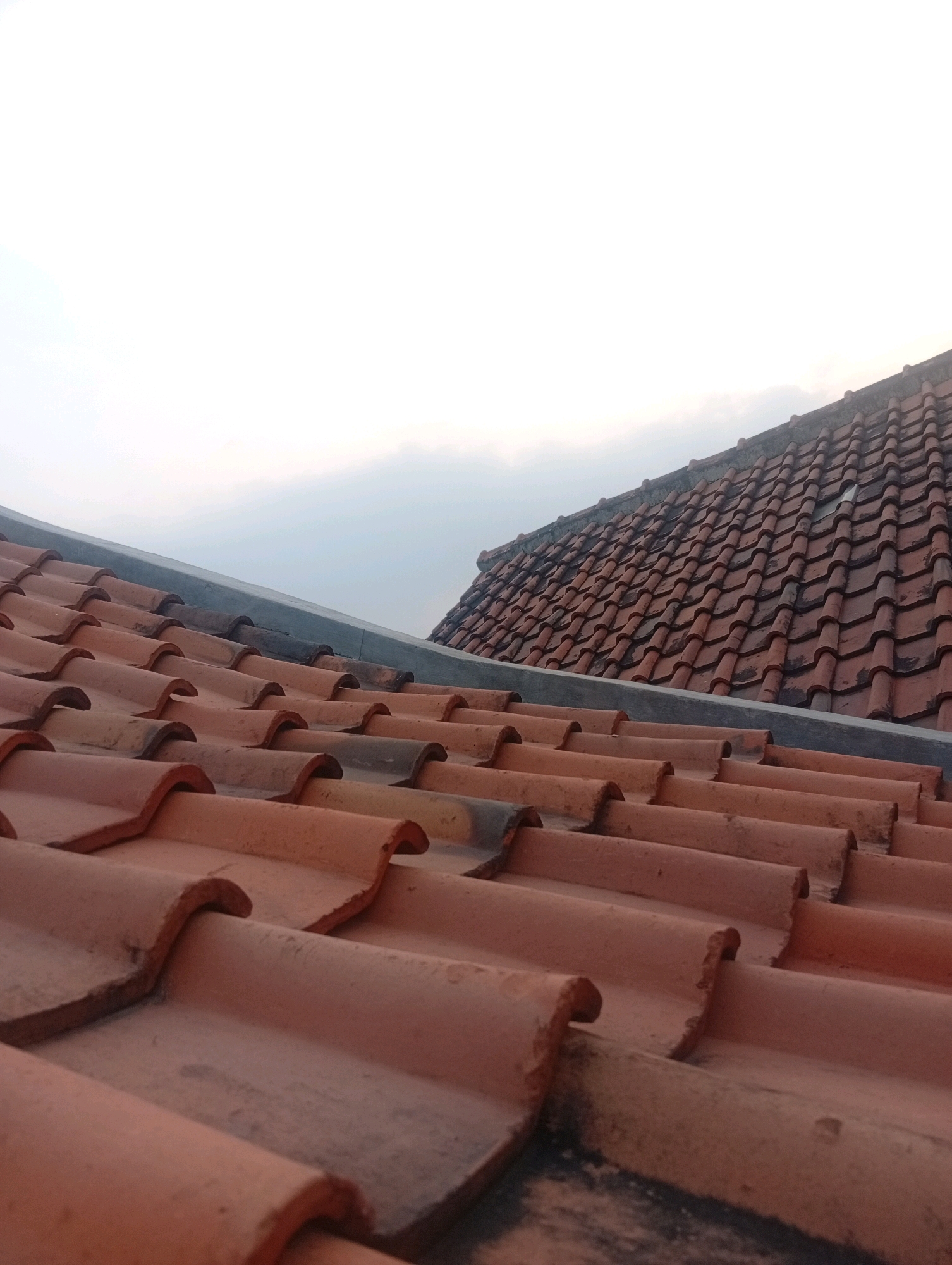 The image shows a close-up view of terracotta roof tiles under an overcast sky, possibly in Kebandingan, Indonesia. The tiles are arranged in overlapping rows, with some showing signs of age and weathering. The perspective is from a low angle, looking up towards the sky, with a second, further roofline visible in the background. No people or actions are depicted in the image. The overall impression is one of a residential or commercial building's rooftop.