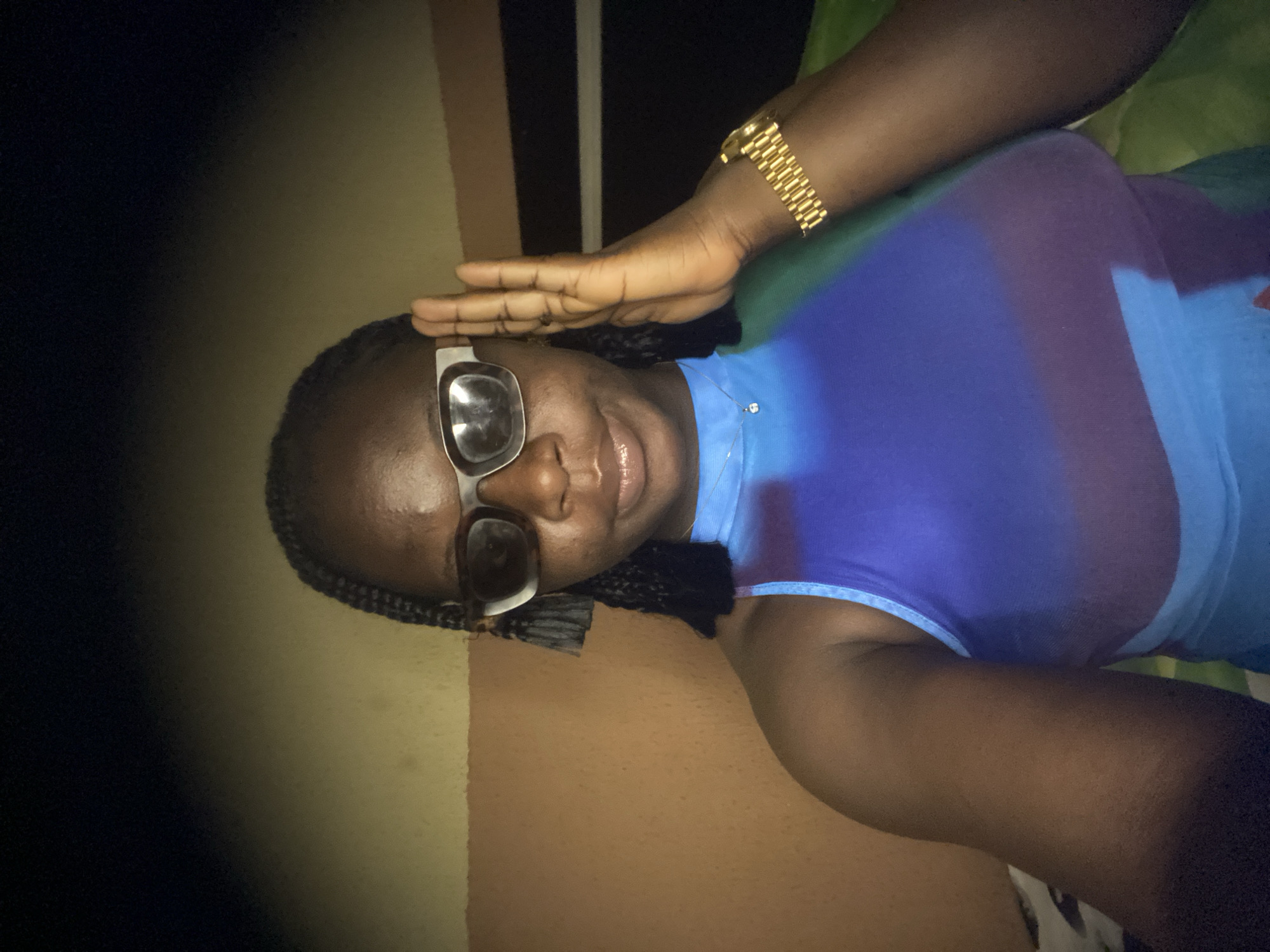 The image shows a person with dark skin and braided hair wearing sunglasses, a blue shirt, and a gold watch. One hand is raised to their forehead. The background appears to be an indoor setting with a wall and furniture. No specific details indicate the location being in the city of Maigizo, Nigeria.