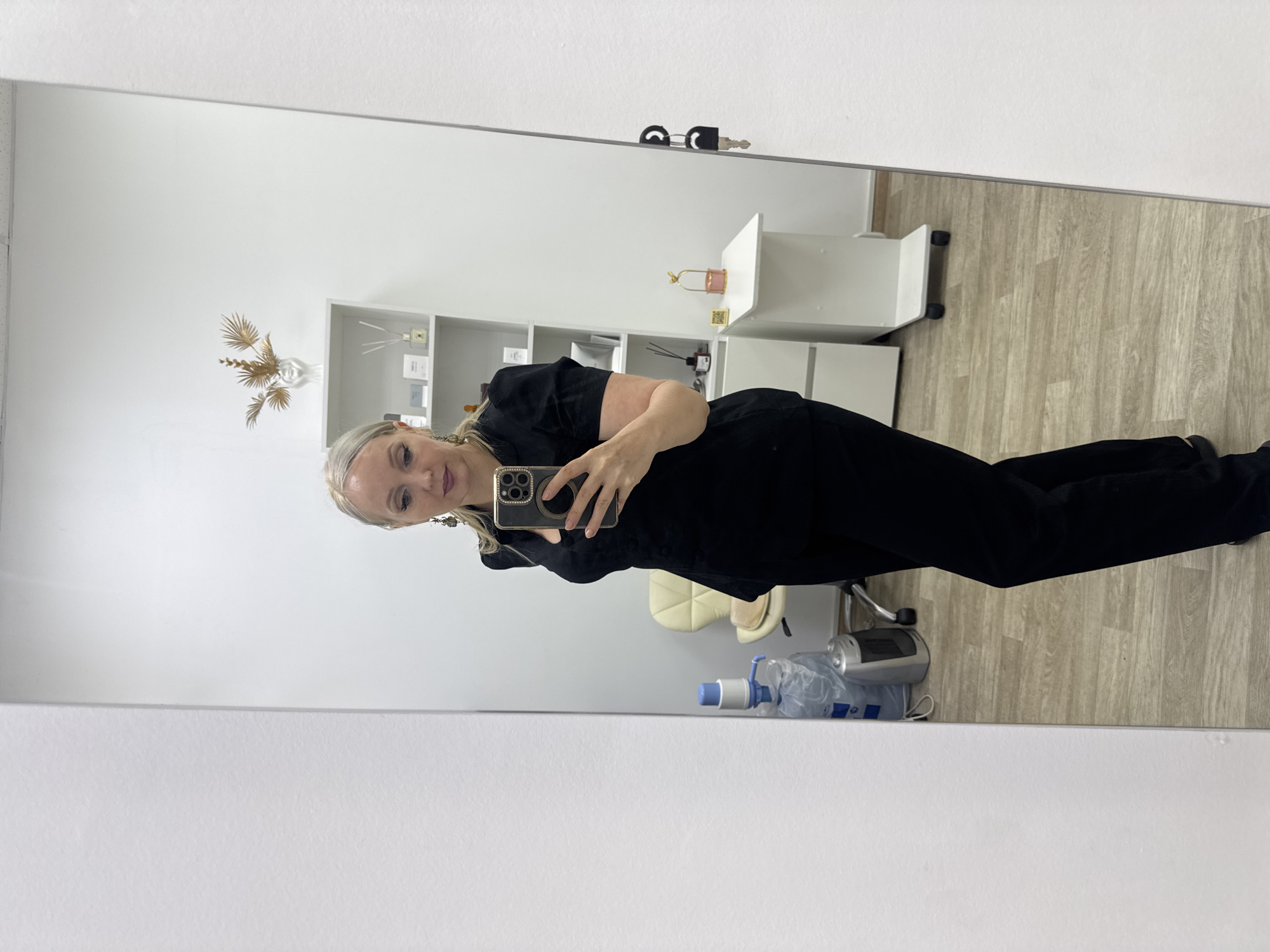 A woman with blonde hair styled in a ponytail takes a selfie in a mirror. She is wearing a black top and black pants. The background shows a room with shelves, a chair, and a small table with decorative items. There is also a water cooler and a small heater visible. The floor is made of light-colored wood. The woman is holding a smartphone in her right hand, which is capturing her reflection. The scene appears to be in an indoor setting, possibly a salon or office. The location is Komsomolsk-on-Amur, Russia.
