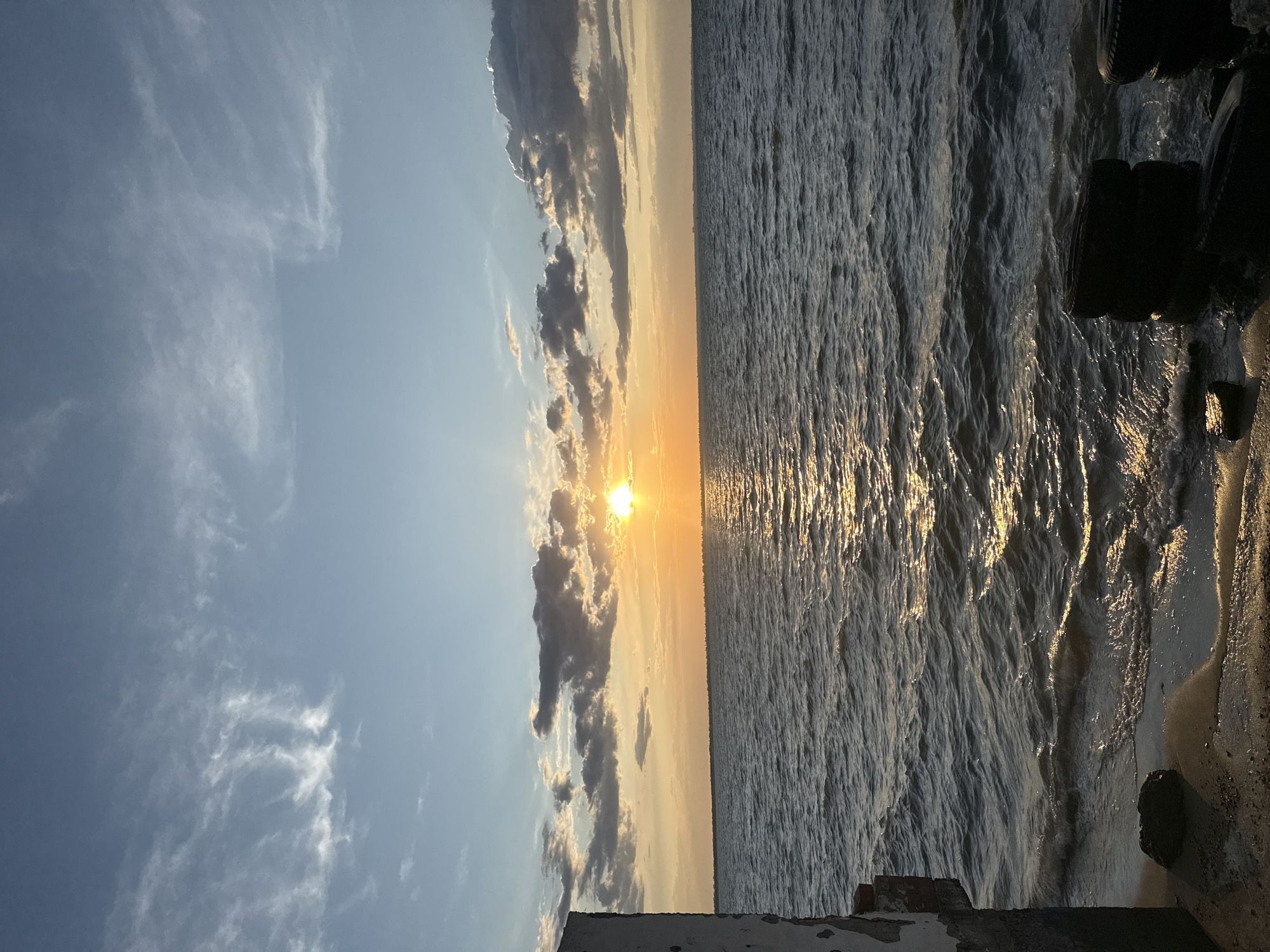 The image shows a wide expanse of choppy, dark blue water under a bright, partly cloudy sky. The sun, a brilliant orange disc, is setting on the horizon, casting a shimmering golden path across the water's surface. Wispy white clouds streak across the upper sky, while darker, heavier clouds are clustered around the setting sun. In the foreground, a concrete structure with weathered white paint and exposed brickwork juts out from the left side of the frame, partially obscuring the shoreline. The shoreline itself is sandy and wet, with a few dark rocks and discarded car tires visible. The overall scene captures a tranquil yet dramatic sunset over the sea, likely near a coastal area.