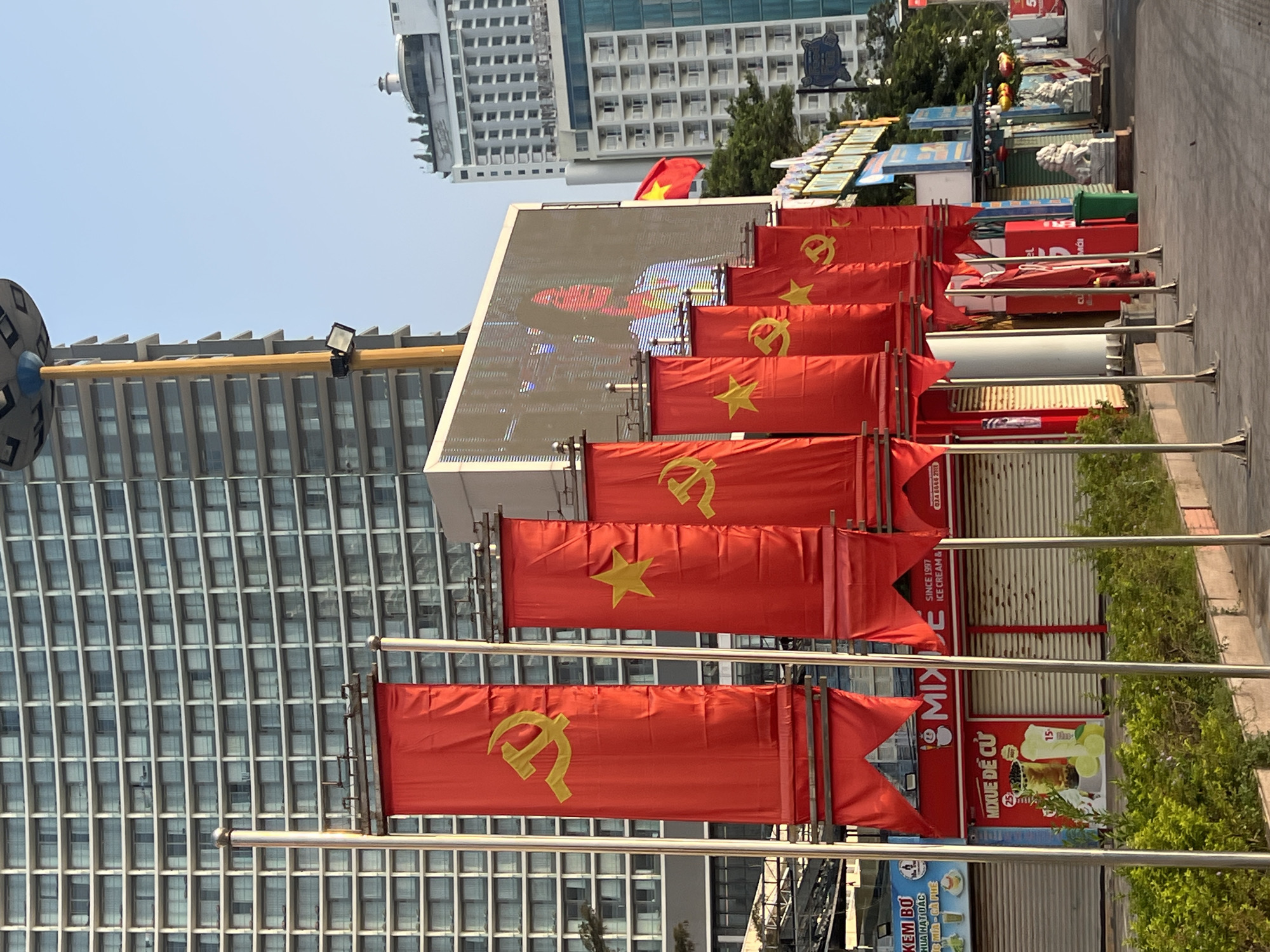 The image captures a street scene in Nha Trang, Vietnam, adorned with communist banners. Red flags, each bearing a golden hammer and sickle, line the street. A large screen displays visuals, while behind the banners, commercial establishments with closed shutters and advertisements are visible. The backdrop features modern high-rise buildings and a clear sky, indicating a daytime setting. The overall scene suggests a public space decorated for an event or commemoration, reflecting the political landscape of Vietnam.