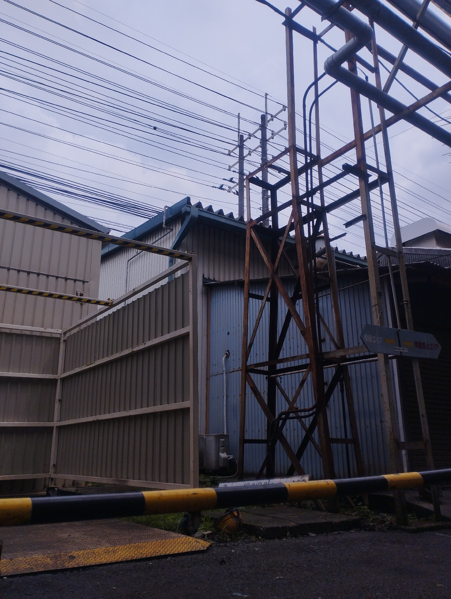 The image, taken in Hamura City, Japan, depicts an industrial area under a cloudy sky. In the foreground, a black and yellow striped barrier arm lies across a textured surface, likely a road or entryway. Behind it, a series of corrugated metal walls, some with yellow and black warning stripes, form a perimeter. A small, blue-grey prefabricated building with a dark blue, sloped roof stands within the fenced area. To the right, a tall, rusty metal frame supports various pipes, and a sign with Japanese text pointing left is visible on one of the support posts. Electrical wires crisscross the sky above the structures, suggesting the presence of power lines or communication cables. The overall scene is one of an operational industrial facility, characterized by utilitarian structures, safety markings, and a functional aesthetic.