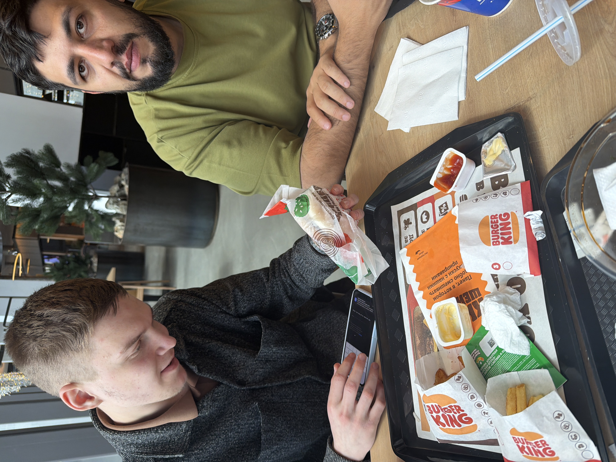Two men are seated at a table in what appears to be a Burger King restaurant. The man on the right is wearing a light green sweater and has a beard. The man on the left is holding a Burger King wrapped sandwich in one hand and a smartphone in the other. The tray in front of him contains a Burger King meal with fries, sauces, and other items. The scene is in Krasnoyarsk, Russia.