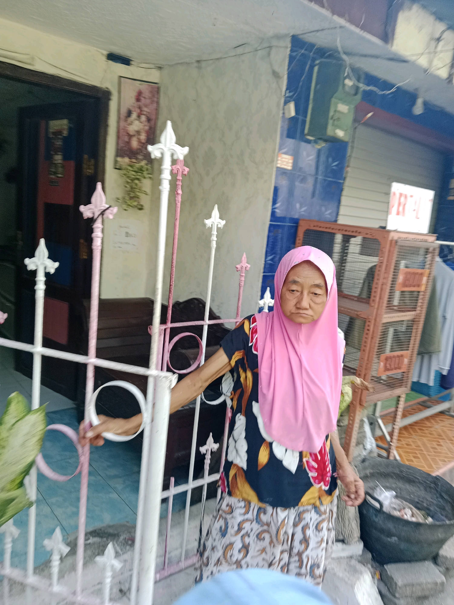 The image shows an elderly woman wearing a pink hijab and a floral patterned shirt, standing behind a white and pink metal gate. She has her right hand on the gate. The background shows a building with blue tiled walls and a metal cage with shelving. Clothes are hanging on the right side of the cage. A large black bin filled with items is visible in the lower right corner. The scene appears to be in a residential area.