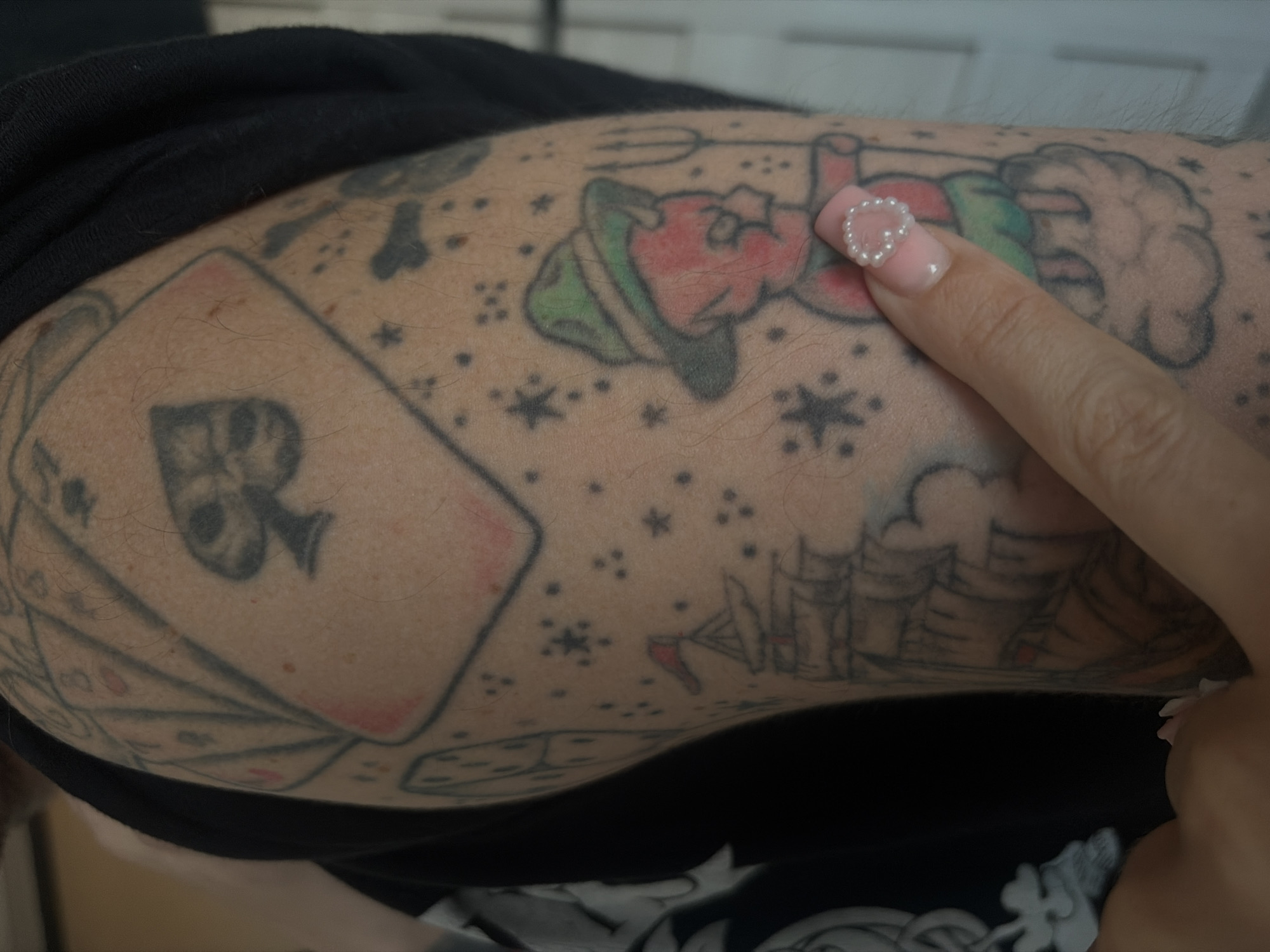 The image shows a close-up of a person's arm, which is covered in tattoos. The tattoos include a large ace of spades card, a sailing ship, dice, and a small red devil figure wearing a green hat. A person's finger, adorned with a pearl-decorated nail, is pointing to the devil tattoo. The background is out of focus and appears to be an indoor setting. The location is not visible in the image, and therefore cannot be specified as Shushary, Russia.