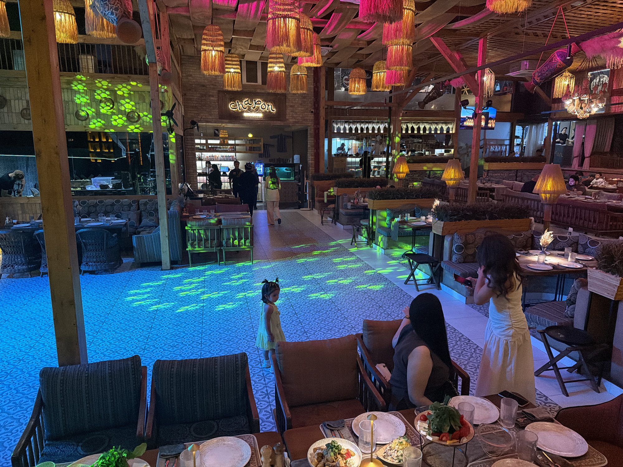 The image depicts the interior of a restaurant in Samarkand, Uzbekistan. The restaurant has a rustic yet modern ambiance, with exposed wooden beams, brick walls, and a variety of seating arrangements, including booth-style seating and individual chairs. The lighting is atmospheric, with colorful lights projected onto the floor and decorative woven lamps hanging from the ceiling. There are several patrons visible in the scene, engaged in dining and socializing. A young girl in a yellow dress is standing on the patterned floor, which is illuminated with green lights. In the foreground, there are tables set with plates, glasses, and food. In the background, a bar area and kitchen can be partially seen, with staff members working. The overall impression is of a vibrant and lively dining establishment.