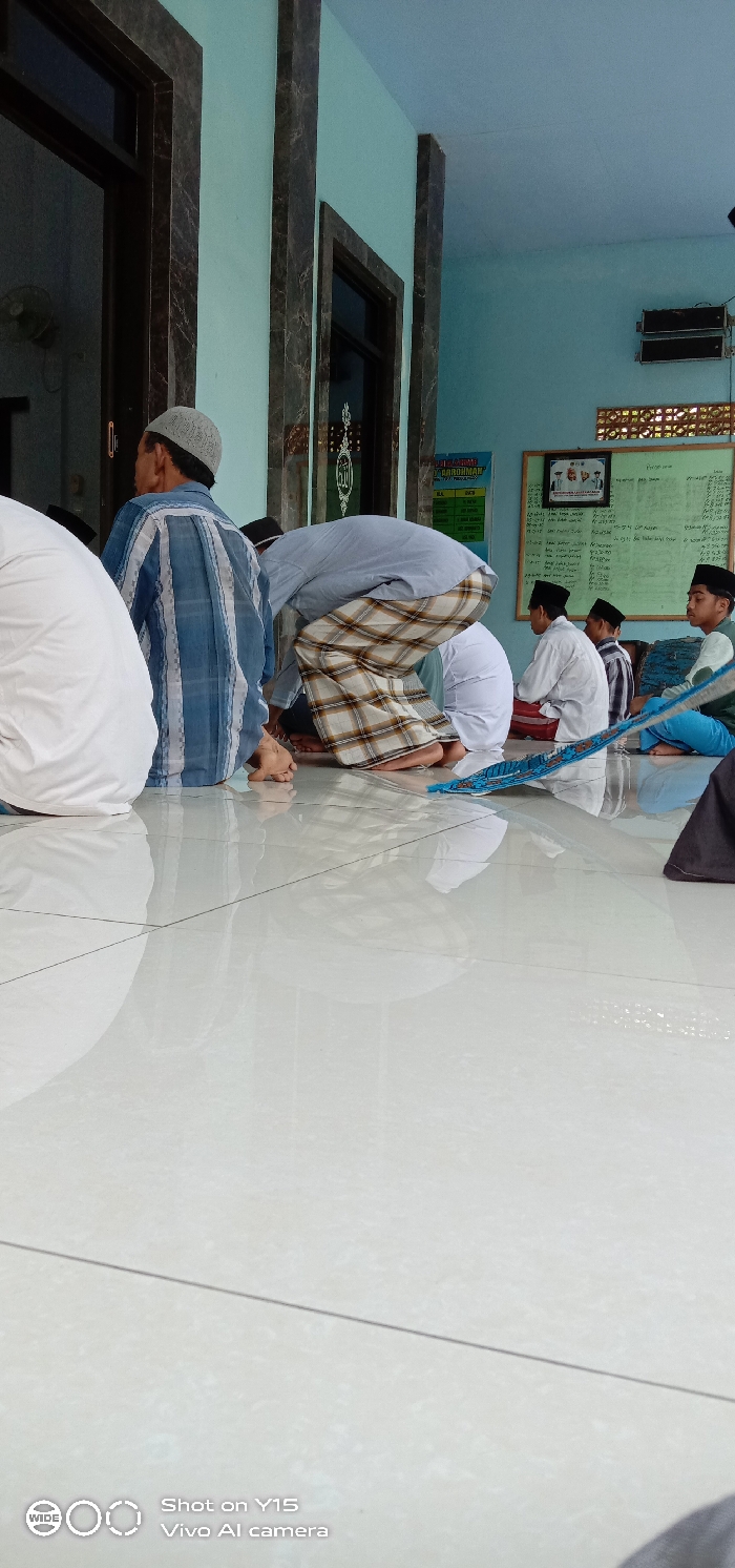 Several people are seated on a shiny white floor inside a room with light blue walls. Most are wearing traditional Islamic clothing. Some face toward the wall, while others sit with their legs crossed. A man in a plaid sarong is bent over in a prayer position. A bulletin board with a schedule or list of names is visible in the background. The room appears to be a prayer area or gathering space.