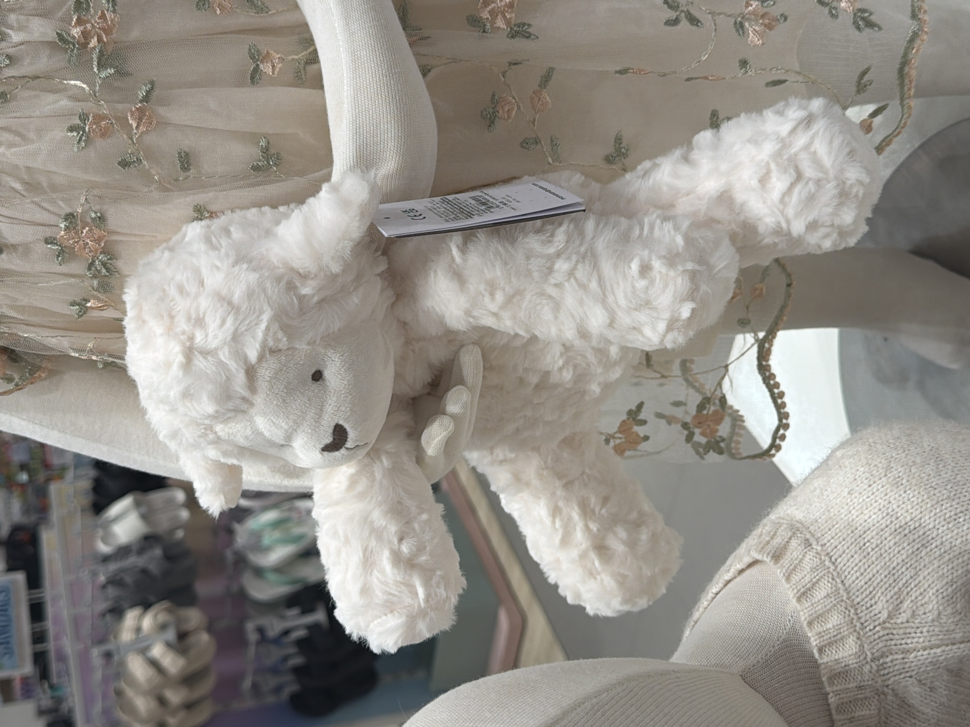 A white, fuzzy stuffed lamb is being held in front of a light-colored mannequin wearing a sheer dress with floral embroidery. The lamb has a small brown nose and dark eyes. A white tag is attached to its arm. In the blurred background, racks of clothing and shoes are visible, suggesting a retail environment. The location is described as Ko Samui District, Thailand.
