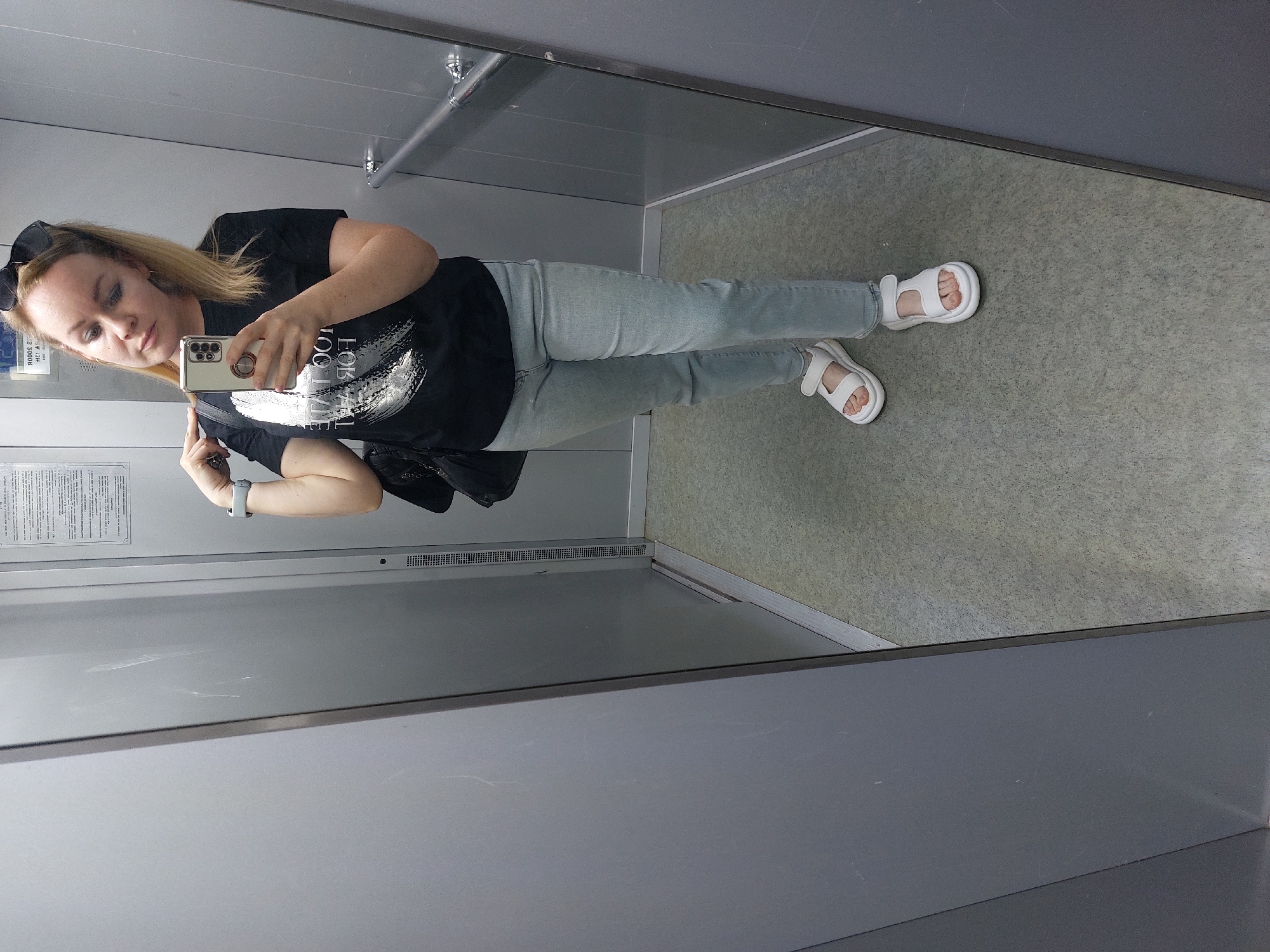 A woman is taking a selfie in an elevator. She is wearing a black t-shirt, light blue jeans, and white sandals. She has sunglasses on top of her head and is holding a smartphone. The elevator has a gray interior. The location is Chaikovsky, Russia.