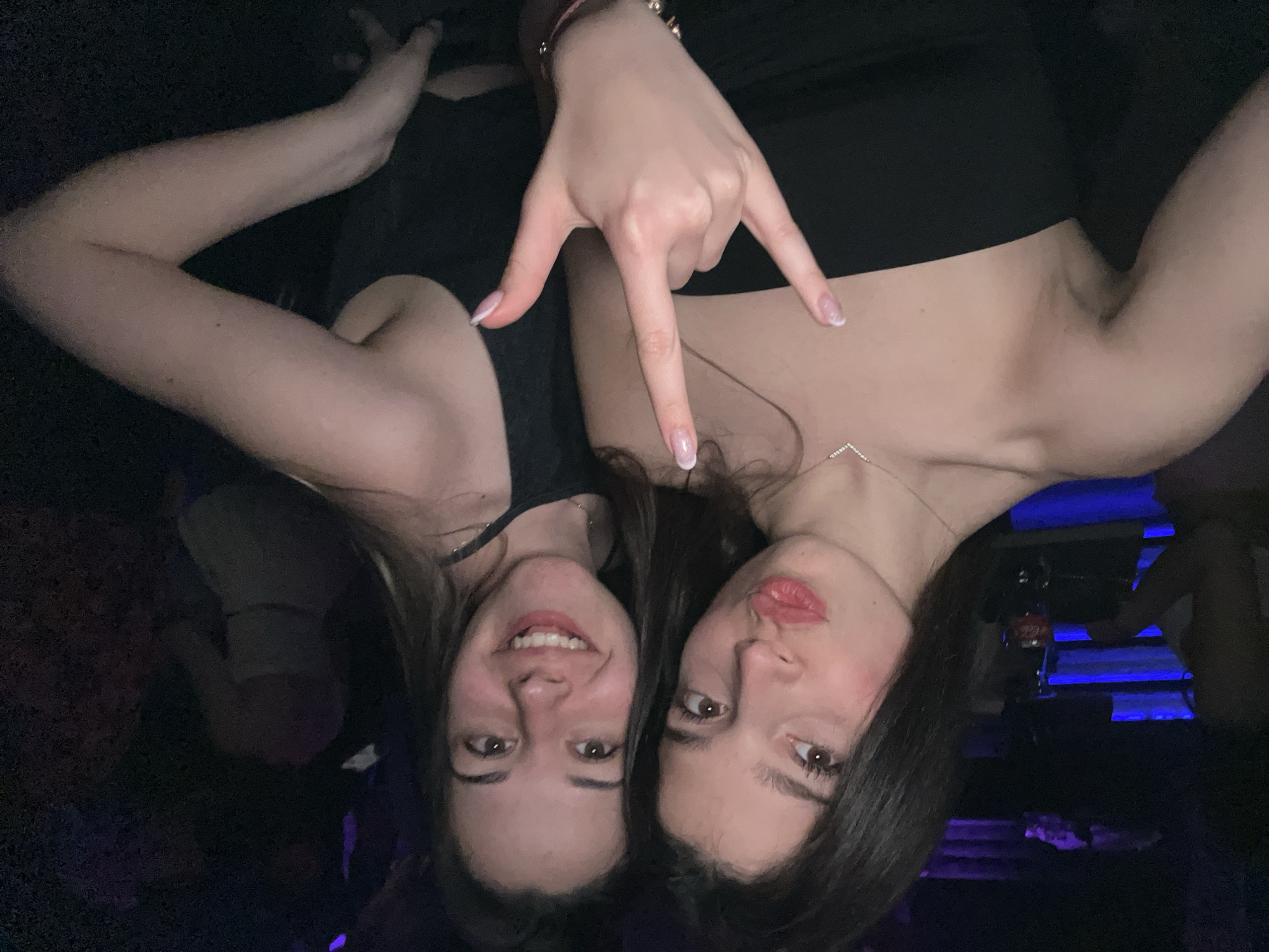 Two young women are posing for a selfie in a dimly lit environment. The woman on the left is smiling, while the woman on the right is pouting. The woman on the right is making a hand gesture with her fingers extended in a "rock on" fashion. They are wearing dark-colored tops. A bar and drinks are visible in the background. The location is likely a nightclub in Varna, Bulgaria.