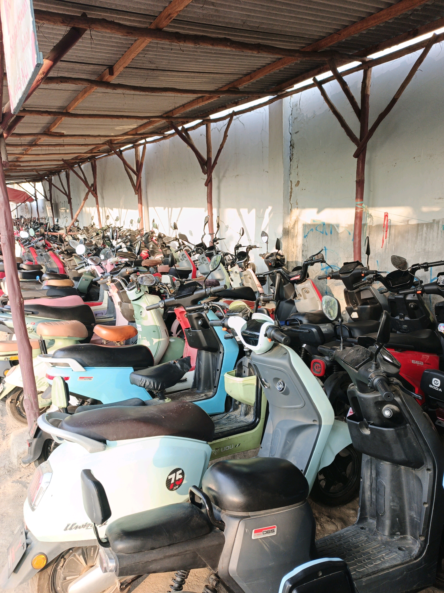 The image depicts a dense outdoor parking area filled with numerous electric scooters and motorcycles, all parked under a makeshift corrugated metal roof supported by rough wooden beams and posts. The scooters are varied in color, with prominent shades including light blue, green, white, black, and red. Many have visible seats, handlebars, mirrors, and wheels. Some scooters feature decals or branding, such as a "75" emblem on a white scooter. The environment appears somewhat weathered, with a light-colored wall in the background showing cracks and graffiti, and the ground is covered with dirt and debris. The lighting suggests it is daytime, with sunlight casting shadows. There are no people visible in the frame. The scene captures a common urban or semi-urban sight in many parts of the world where personal electric transportation is prevalent, and it is plausible to find such a parking arrangement in a location like Krimun, Indonesia, given the prevalence of scooters in Southeast Asia and the typical construction of such shelters in tropical climates.