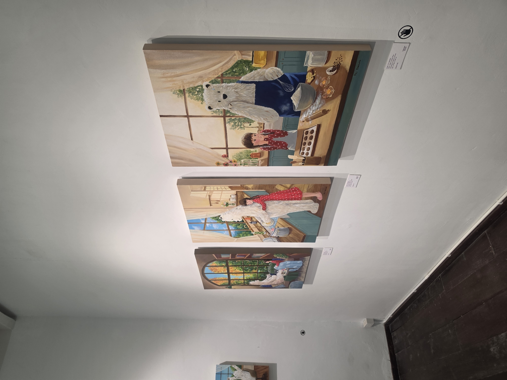 The image shows three paintings hanging on a white wall in what appears to be a gallery or exhibition space. The paintings are arranged vertically. The top painting depicts a child and a large, fluffy white bear wearing a blue apron in a kitchen setting, seemingly baking or cooking together. The middle painting portrays a child in a red dress lying on top of the same white bear in a room with a window. The bottom painting shows the child and the bear near a window with an autumn view.
To the right of the top painting is a sign with an image of a hand inside a circle, and text below it.
The floor is dark wood.
Without further information, the precise location within Bangkok, Thailand is not discernible.