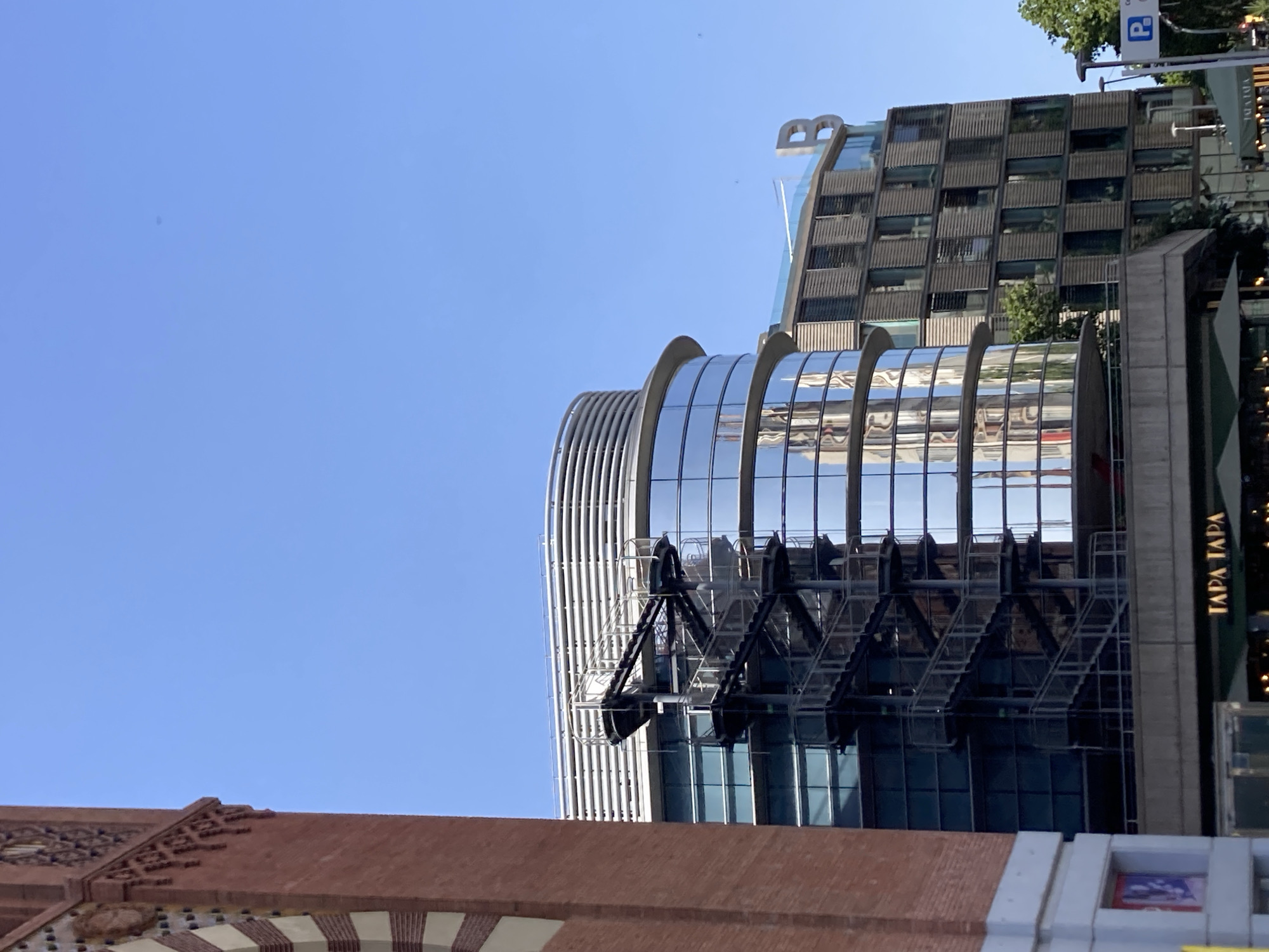 The image captures a street scene in Barcelona, Spain, dominated by modern architecture under a clear blue sky. On the left, a portion of a red brick building, possibly a historic structure with decorative elements, is visible. Adjacent to it stands a contemporary glass-fronted building with a curved facade, reflecting the surrounding environment, including what appears to be a spiral staircase on its exterior. To the right, another building with a grid-like pattern of windows and louvers is seen, topped with a large, stylized letter "B." Below these buildings, a street-level view shows awnings and signage, with "TAPA TAPA" prominently displayed, indicating a restaurant or bar. A blue parking sign with a white "P" is also visible in the distance. The overall impression is one of urban development, blending older architectural styles with newer, sleek designs.
