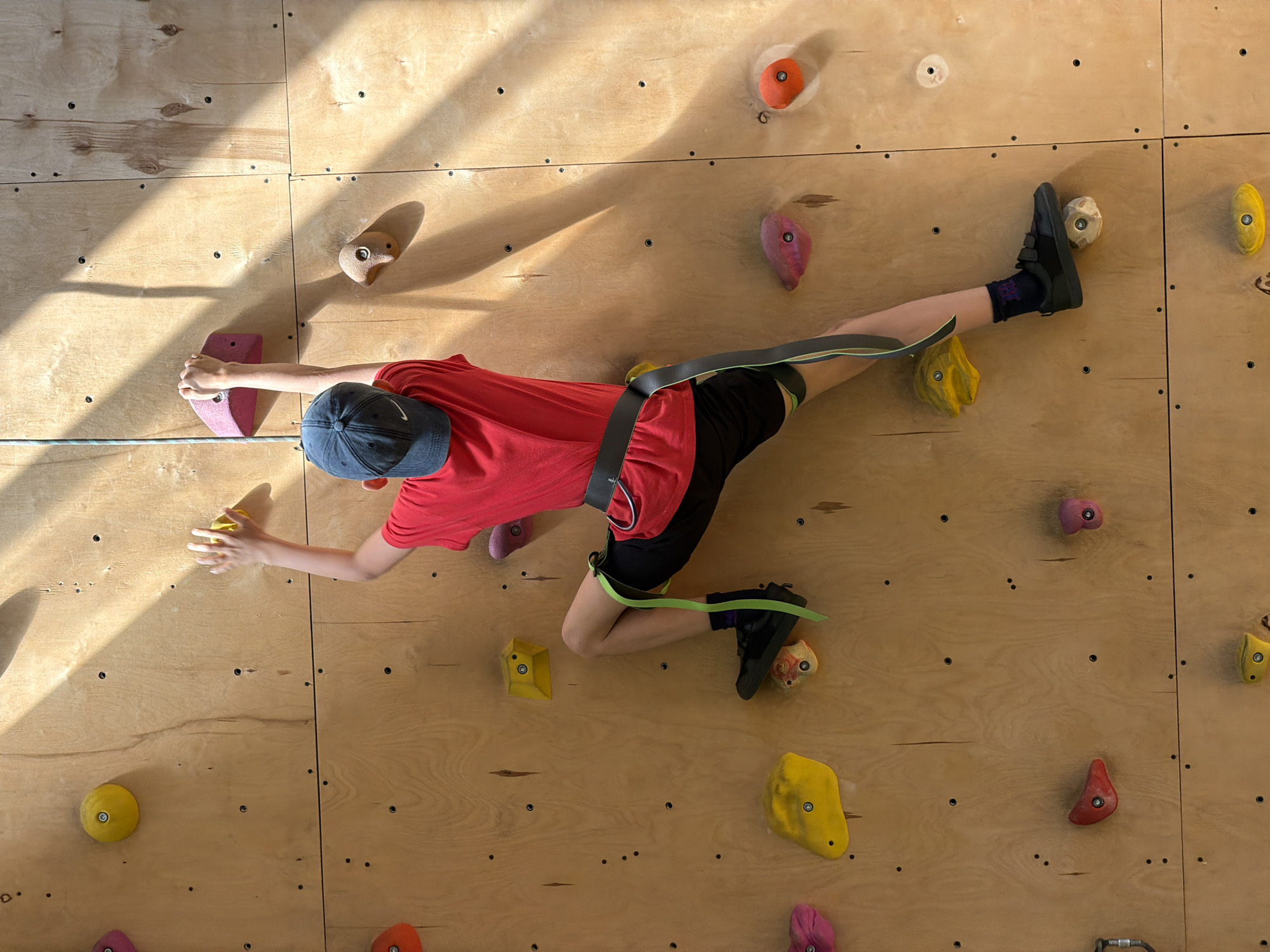 A person is rock climbing on an indoor wall constructed of wood panels with attached colorful climbing holds. The person is wearing a red shirt, black shorts, a blue baseball cap, climbing shoes, and a climbing harness with a rope attached. The climbing wall is located in an unidentified indoor facility. The image was taken in Izhevsk, Russia.