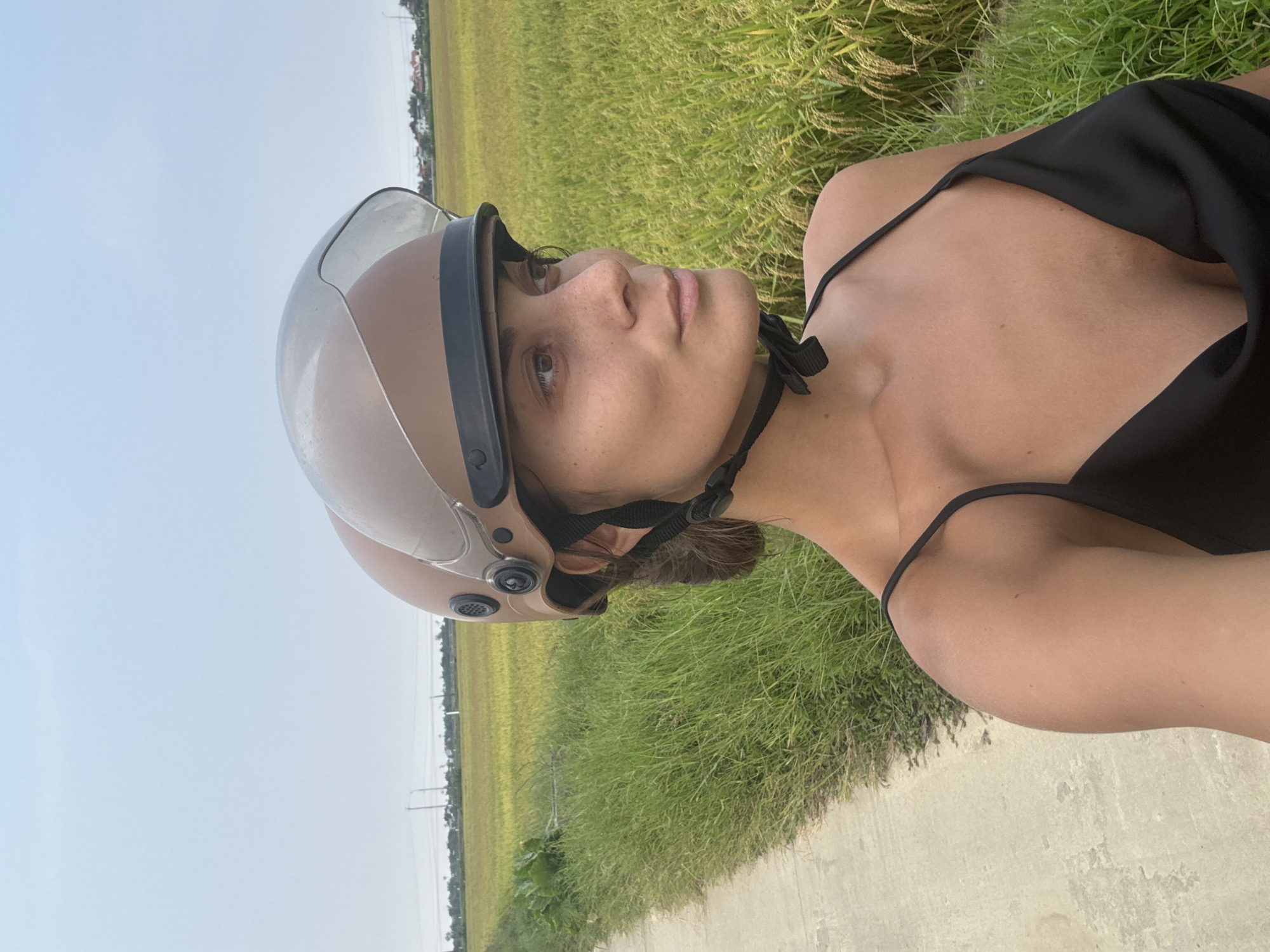 A close-up, portrait-oriented shot features a person, likely female, captured from the chest up. She is wearing a light brown or beige open-face helmet with a clear visor, secured by a black chin strap. Her dark hair is pulled back, visible beneath the helmet. She wears a black sleeveless top with thin straps, revealing her bare shoulders and collarbone. Her gaze is directed upwards and to the right, with a calm and contemplative expression.
The setting appears to be a rural or semi-rural landscape, indicative of Da Nang, Vietnam. Behind the person, a vast expanse of green and golden-yellow crops, characteristic of rice paddies, stretches into the distance. A narrow, light-colored concrete or dirt path runs along the edge of the field, separating it from the viewer. In the far background, on the horizon, faint outlines of buildings or houses and utility poles can be seen against a clear, pale blue sky. The bright, even lighting suggests it is daytime, possibly late morning or afternoon, under clear weather conditions. The presence of the helmet implies the person is on or near a scooter or motorcycle, a common mode of transportation in the region, perhaps taking a break to observe the scenery. There is no visible text in the image.