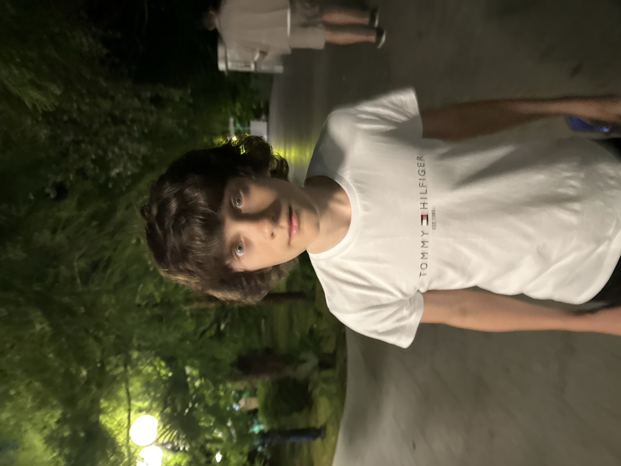 The media file is an image of a young person, likely male, standing outdoors at night. The subject is looking towards the camera with a neutral expression. They are wearing a white t-shirt with the "TOMMY HILFIGER EST. 1985" logo visible on the left side of the chest. The background is dark, with blurred green foliage suggesting trees or bushes. There are also some bright, out-of-focus lights, possibly streetlights or lamps, contributing to the nighttime setting. Another person is visible in the background, also wearing white clothing, and appears to be walking away. The ground appears to be a paved path or sidewalk. The overall mood is casual and candid, typical of a personal photograph taken during an evening outing. The location context is Sochi, Russia.