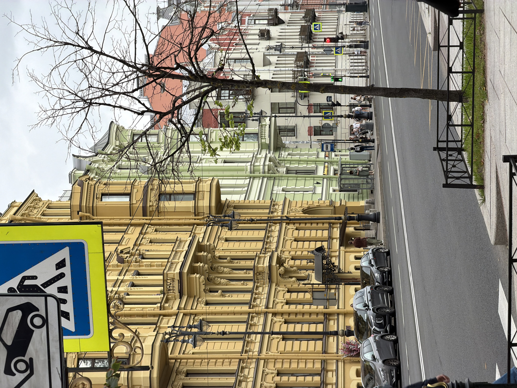 This image shows a street scene in Saint Petersburg, Russia, on a cloudy day. The foreground features a sidewalk with a black metal fence enclosing a small patch of grass and a tree. Across the street, a row of historic buildings with ornate facades in yellow, green, and red lines the street. There are several cars parked along the curb, including a gray SUV and a black sedan. Pedestrians are visible on the sidewalk and crossing the street. Traffic lights and street signs, including a pedestrian crossing sign, are also present. The overall impression is of a busy urban environment with a blend of modern and historical architecture.