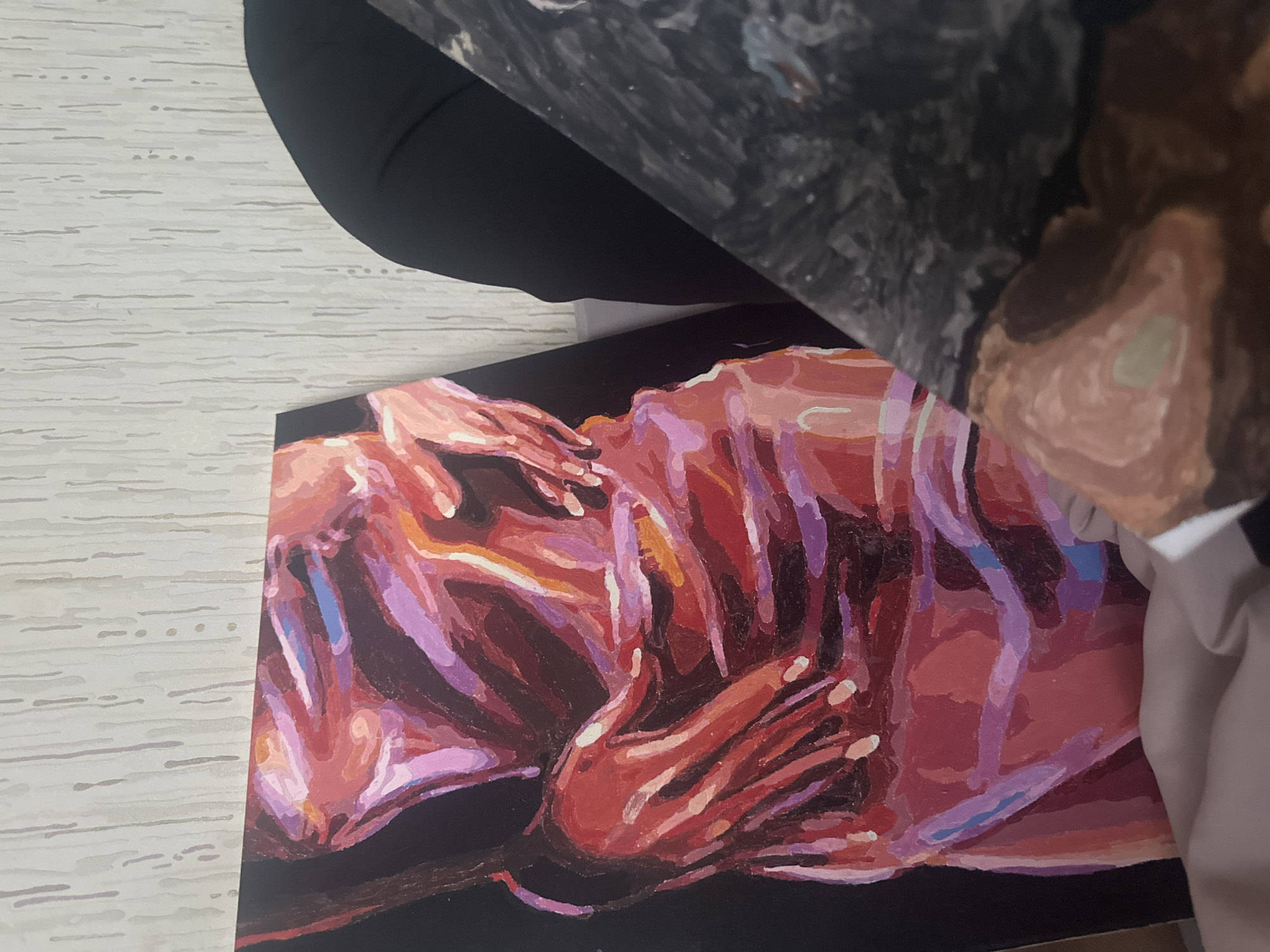 The image shows a painting lying on a surface that appears to be light-colored wood grain. The painting features two figures in a stylized, somewhat abstract manner, rendered in shades of red, pink, purple, and brown. The figures are intertwined and appear to be lying down, with hands prominently displayed. The background of the painting is black. Parts of other objects are present in the image, including an expanse of dark fabric and a section with shades of gray, brown, and white. The image does not provide enough information to determine the location or any details regarding the city of Высокий, Russia.