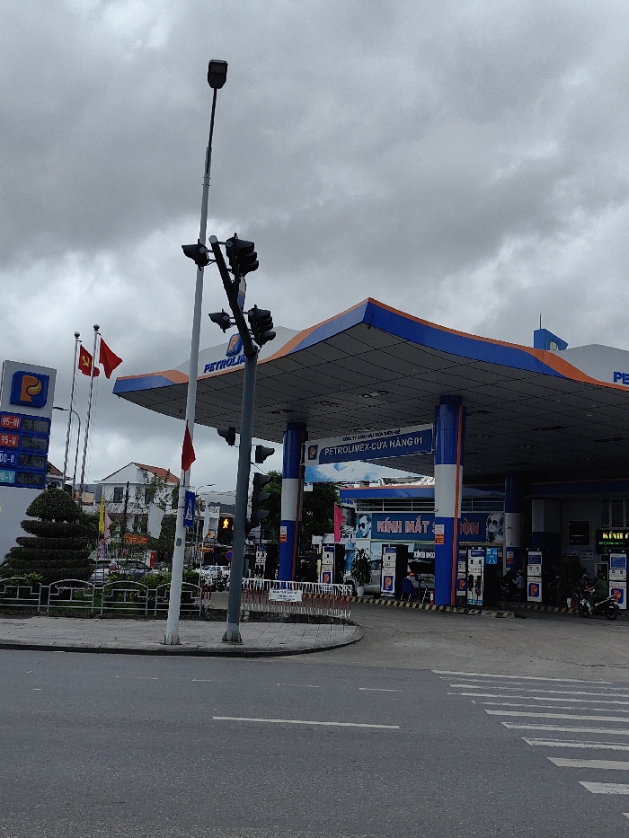 The image depicts a Petrolimex gas station situated at a street corner under an overcast sky in Thuận Hóa Ward, Vietnam.
The Petrolimex station features a large canopy with a prominent blue and orange trim, supported by several blue pillars. A white sign on the canopy identifies it as "CÔNG TY XĂNG DẦU THỪA THIÊN HUẾ PETROLIMEX-CỬA HÀNG 01" (Thua Thien Hue Petroleum Company Petrolimex - Store 01). To the left, a tall sign displays the Petrolimex logo and digital prices for various fuel types, including "95-V," "95-IV," and "DO-V." Two flagpoles stand near this sign, flying the red Vietnamese national flag with a yellow star and a solid red flag. Under the canopy, multiple fuel pumps are visible, alongside several parked motorbikes and a few individuals; one person appears to be fueling a motorbike. Behind the pumps, part of a building is visible with "KÍNH MẮT" (eyeglasses) signage.
In the foreground, an asphalt road includes a solid white line and a designated pedestrian crossing with white zebra stripes. A concrete sidewalk curves around a small, fenced, triangular median strip that contains a manicured, tiered green bush. Two tall poles stand at the intersection: one a street lamp, the other supporting multiple clusters of traffic lights. A small red flag is attached to one of these poles. In the background, across the street from the gas station, various multi-story buildings and additional vehicles are visible. The sky is uniformly gray and overcast.