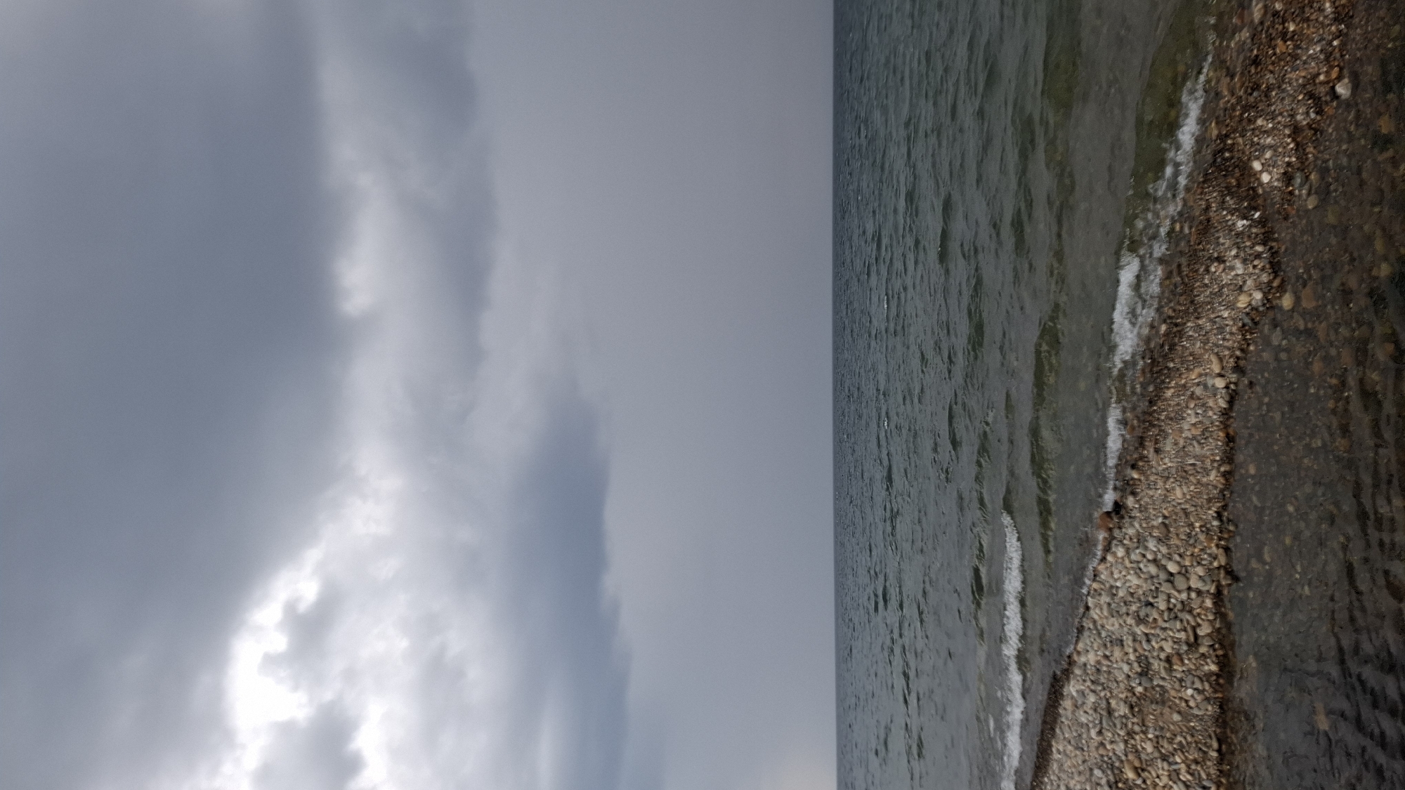 The image depicts a moody, overcast scene at the shore of a large body of water, possibly Lake Baikal, near the city of Priboi, Russia. The sky is filled with heavy, grey clouds, suggesting an impending storm or recent rainfall. The water is a deep, dark blue-green, with visible ripples and small waves lapping against the shore. The shoreline is composed of smooth, multicolored pebbles and stones, with a narrow strip of darker, wet sand or mud closer to the water's edge. The overall atmosphere is somber and natural, with no signs of human presence or activity.